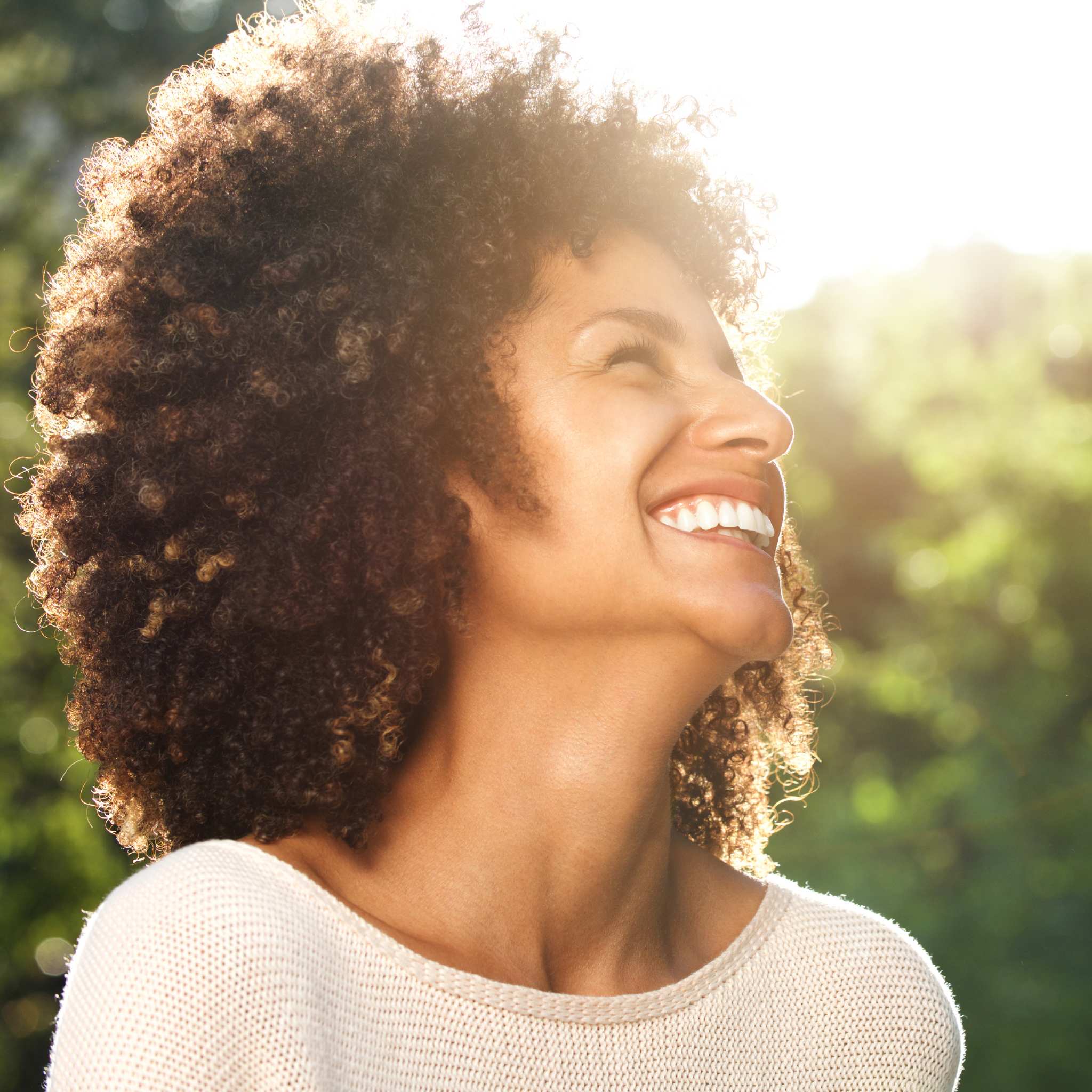 A close-up, outdoor portrait of a joyful woman with curly brown hair and a wide, radiant smile, tilting her head back with her eyes closed in apparent bliss. The sun's bright rays are visible as a lens flare behind her head and shoulders, suggesting warmth, happiness, and the positive energy of the lightworker path alignment dedication.