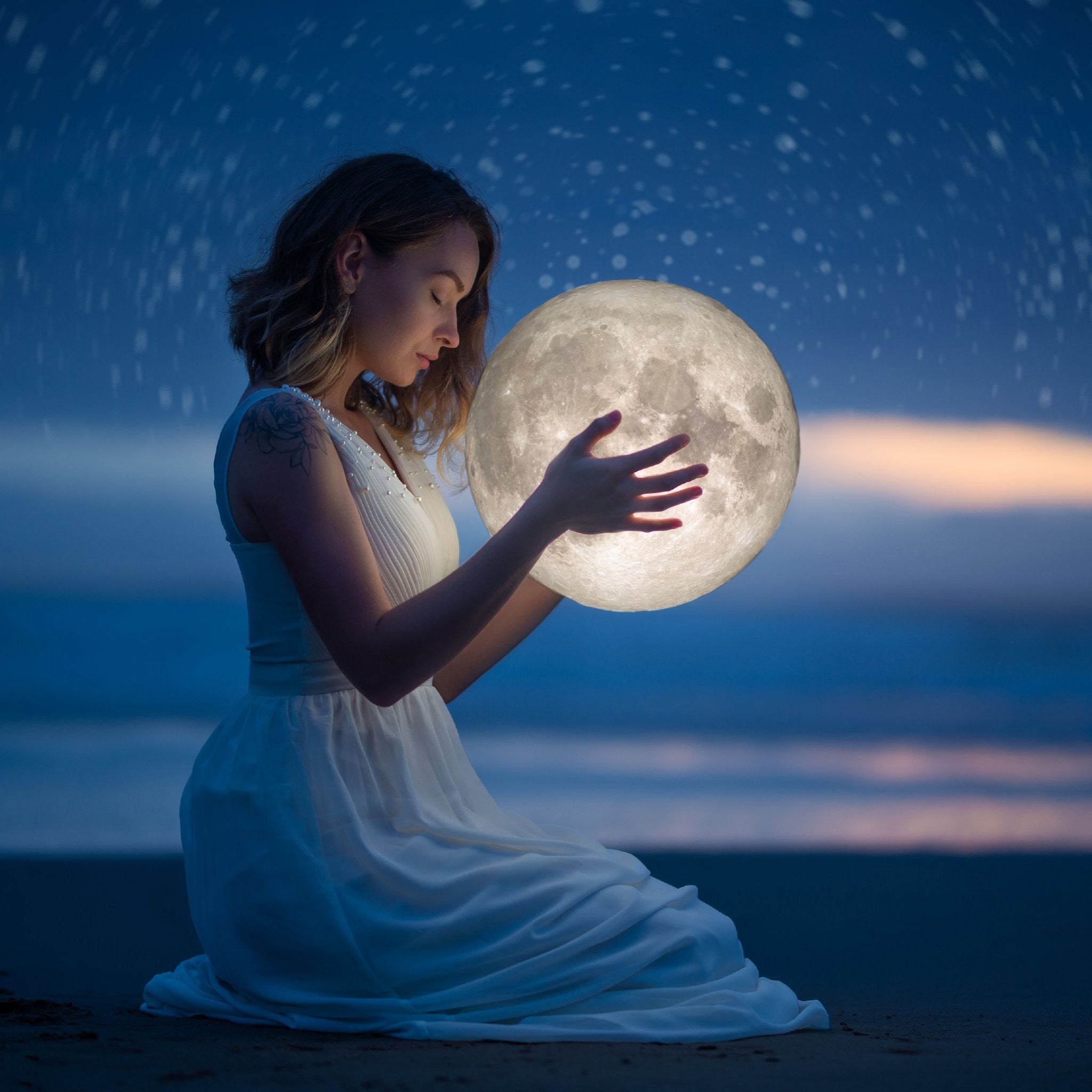 A woman in a white dress kneels on a beach at dusk, holding a large, glowing, illuminated moon sphere in her hands against a dark blue sky with visible stars and a faint horizon.