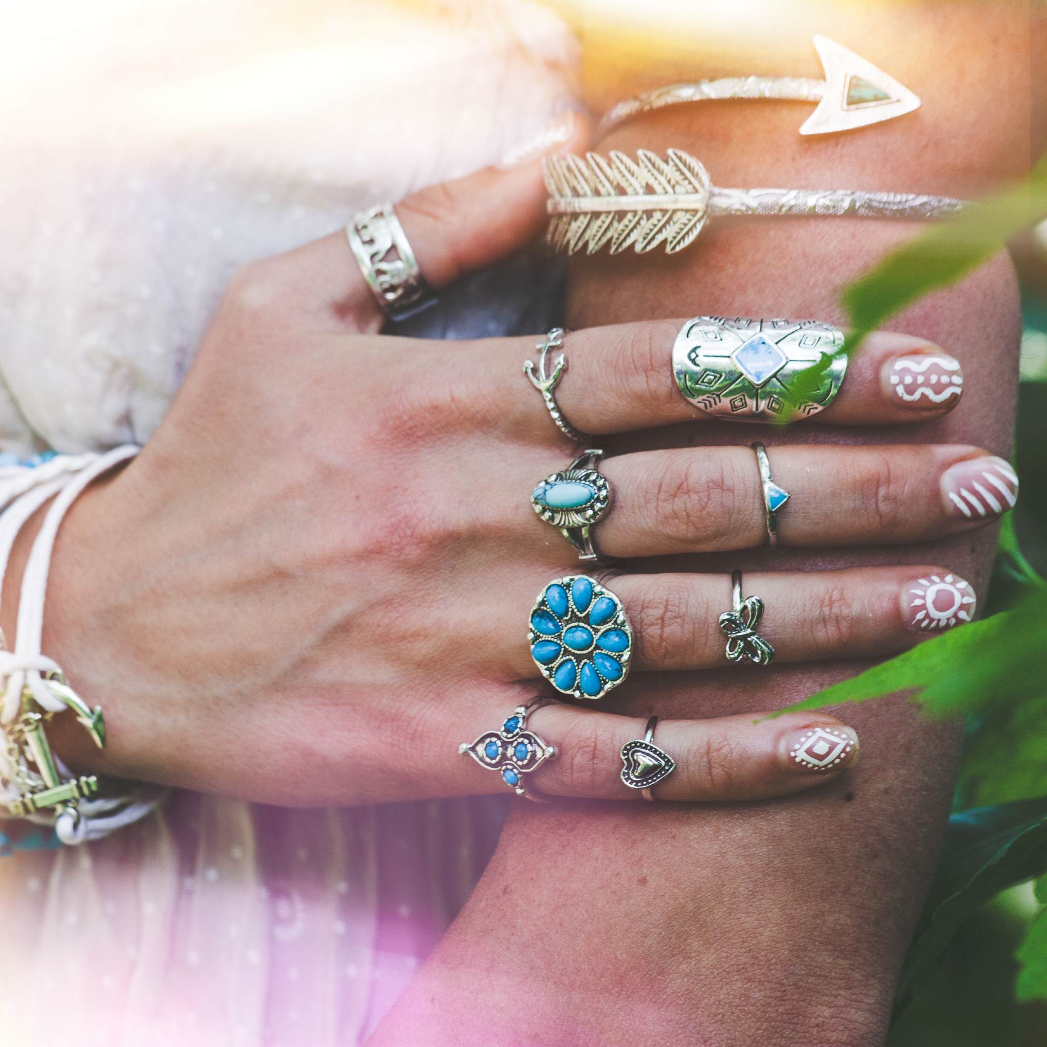 A close-up of a person's arms and hands resting on one another, covered in multiple intricate silver rings with turquoise stones and several silver bracelets, including a feather motif, set against a background of soft green foliage and sunlight.