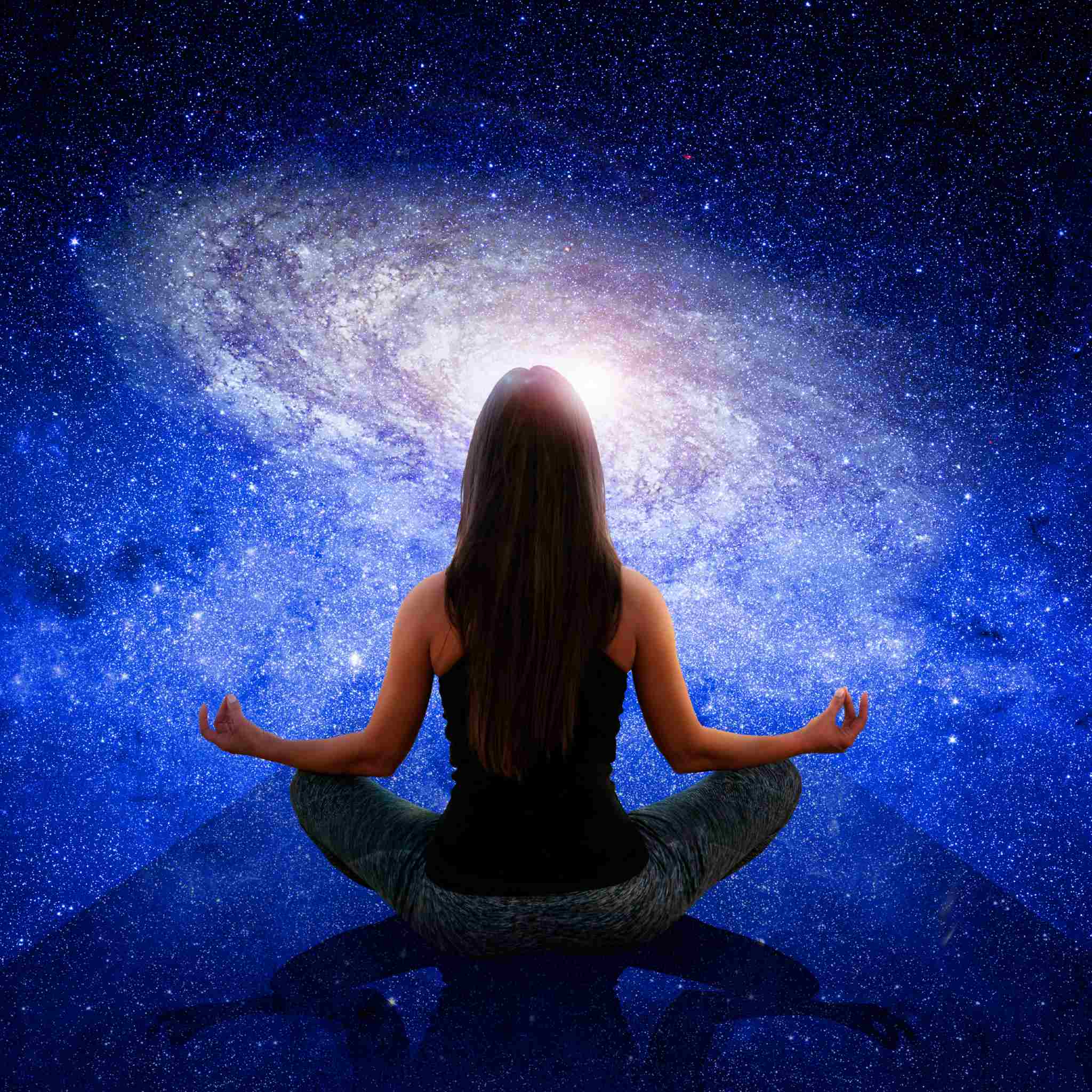 A woman with long dark hair sits cross-legged, viewed from behind, meditating with hands on her knees in a yoga position. Above her head is a large, glowing spiral galaxy set against a dark blue, starry cosmic background, suggesting a cosmic connection.