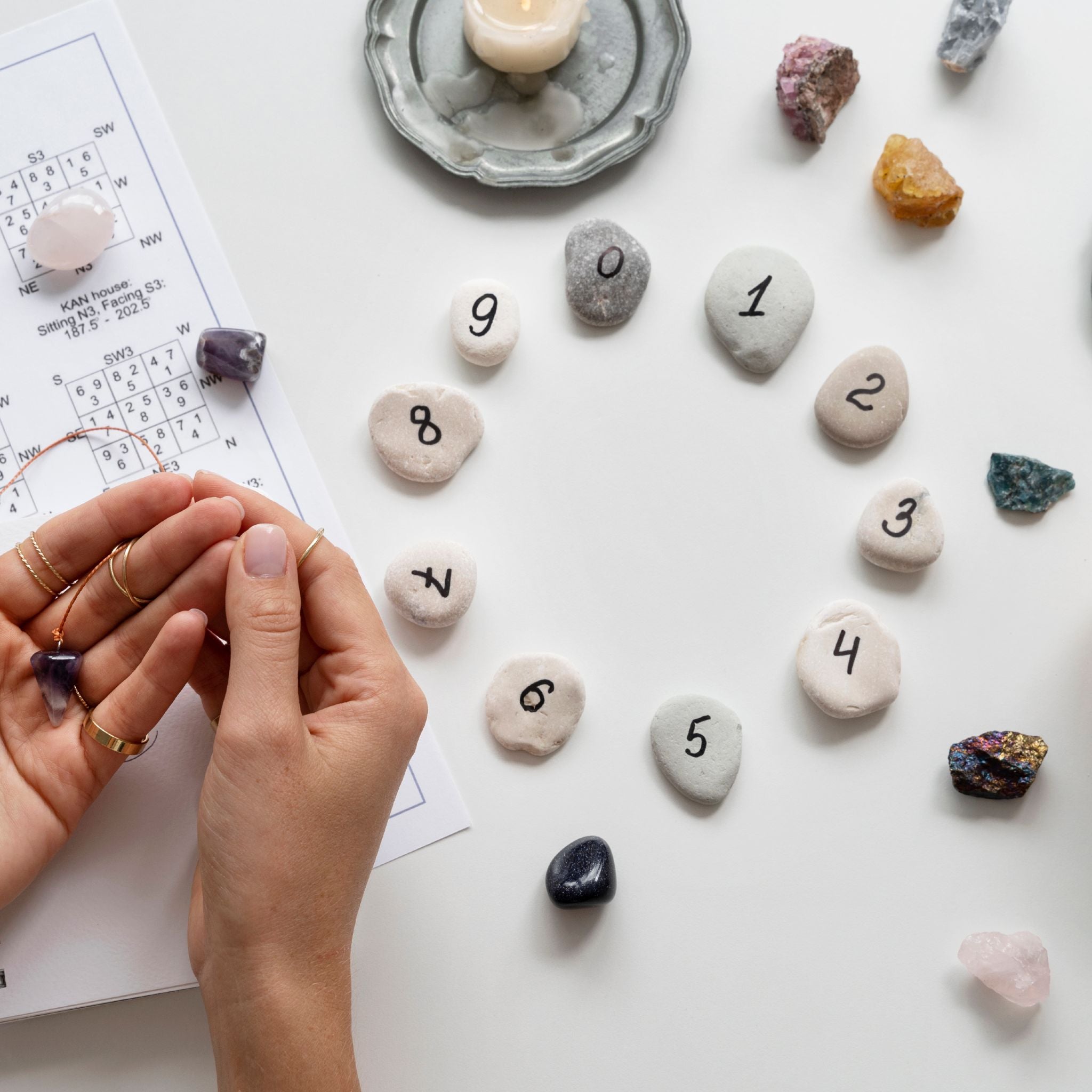 An overhead photograph of a white table showcasing a numerology setup, with small, smooth stones arranged in a circle, each painted with a digit from 1 to 9. The scene also includes various raw and tumbled crystals, a burning tea light candle, and a printed chart, symbolising the spiritual and mathematical focus of the numerology path alignment dedication.