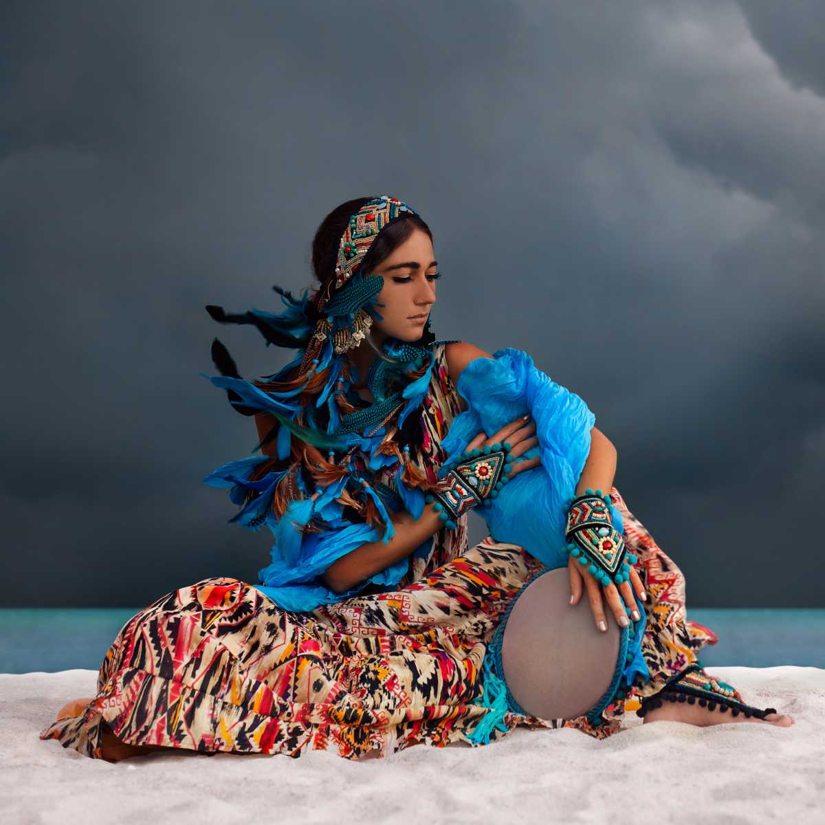 A woman seated on white sand against a dark stormy sky, wearing a colourful patterned dress, bright blue top, matching headpiece with feathers, and patterned arm cuffs while holding a drum.