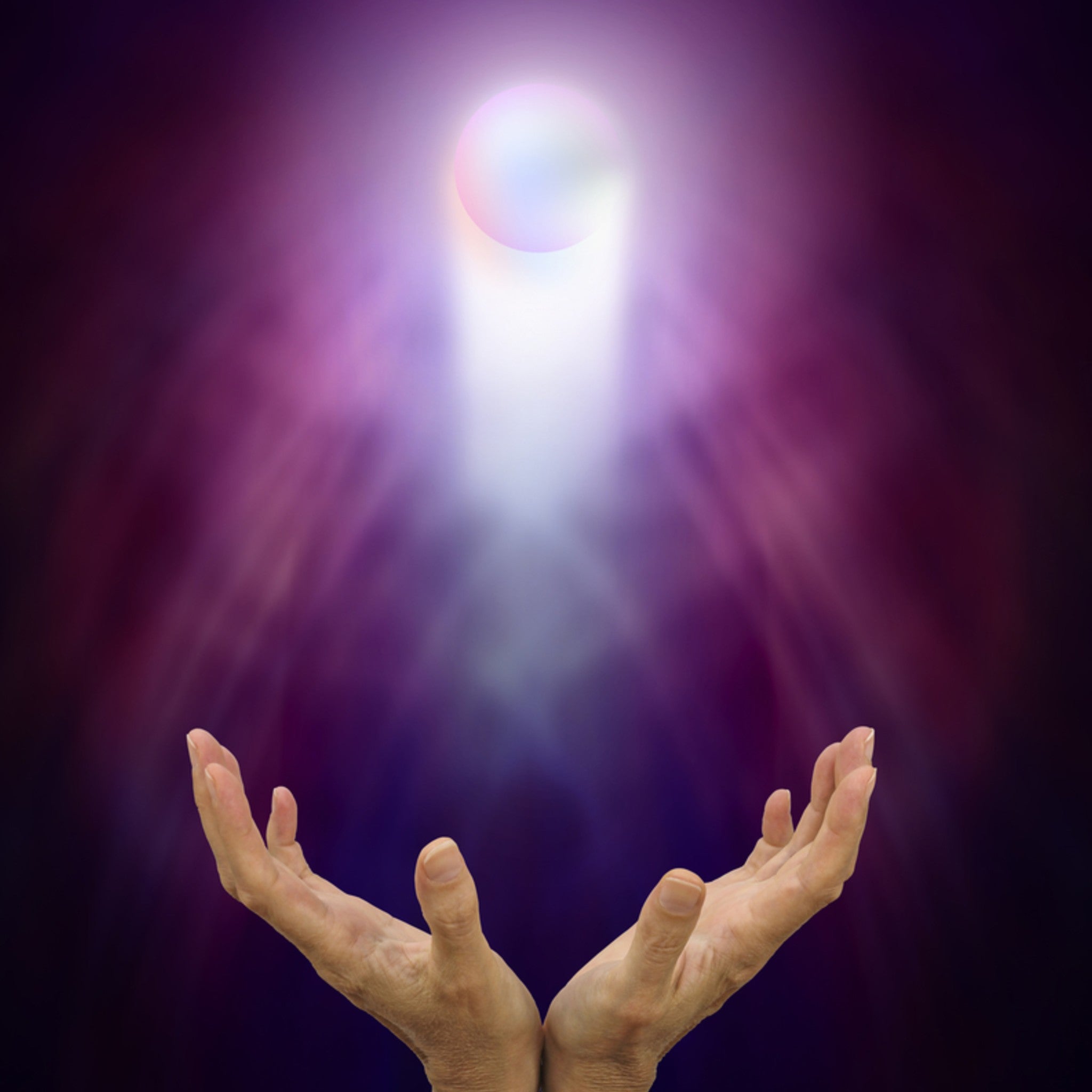 A photograph of a pair of hands, cupped upward, receiving a beam of brilliant white light that streams down from a glowing orb above, against a dark purple, ethereal background. The image symbolises the channelling of Reiki energy for psychic development.