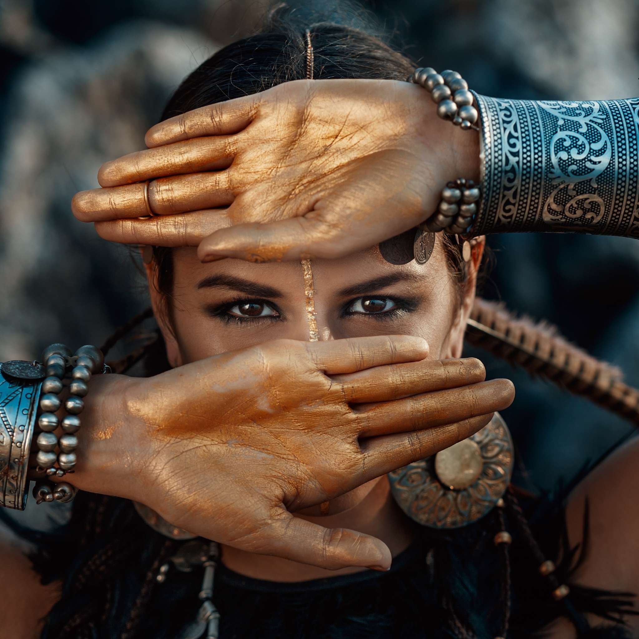 Close-up of a person with tribal-style jewellery, whose hands are painted gold and crossed over their face, allowing only their intense eyes to be seen.