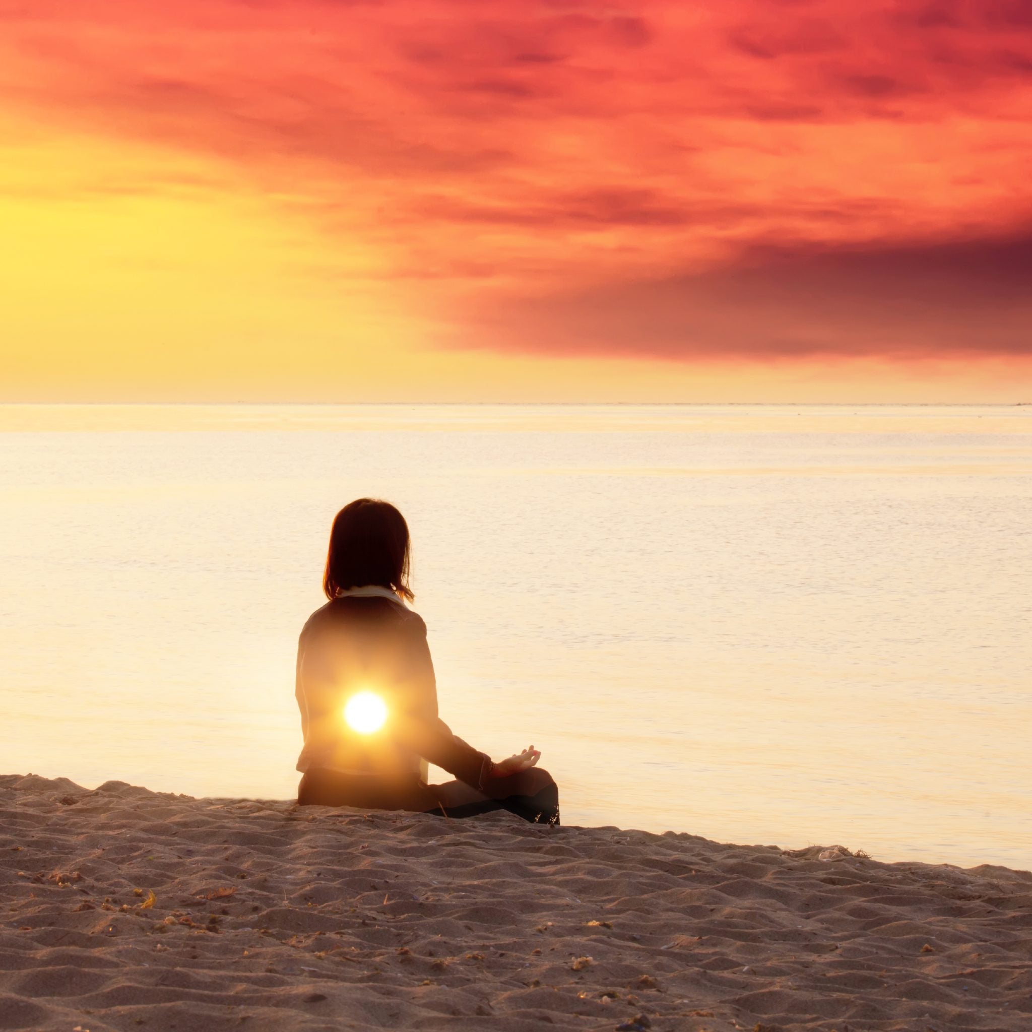 A silhouette of a person sitting cross-legged on a sandy beach, facing a stunning sunset over the ocean, with an intense, glowing yellow sunburst light emanating from their solar plexus area.