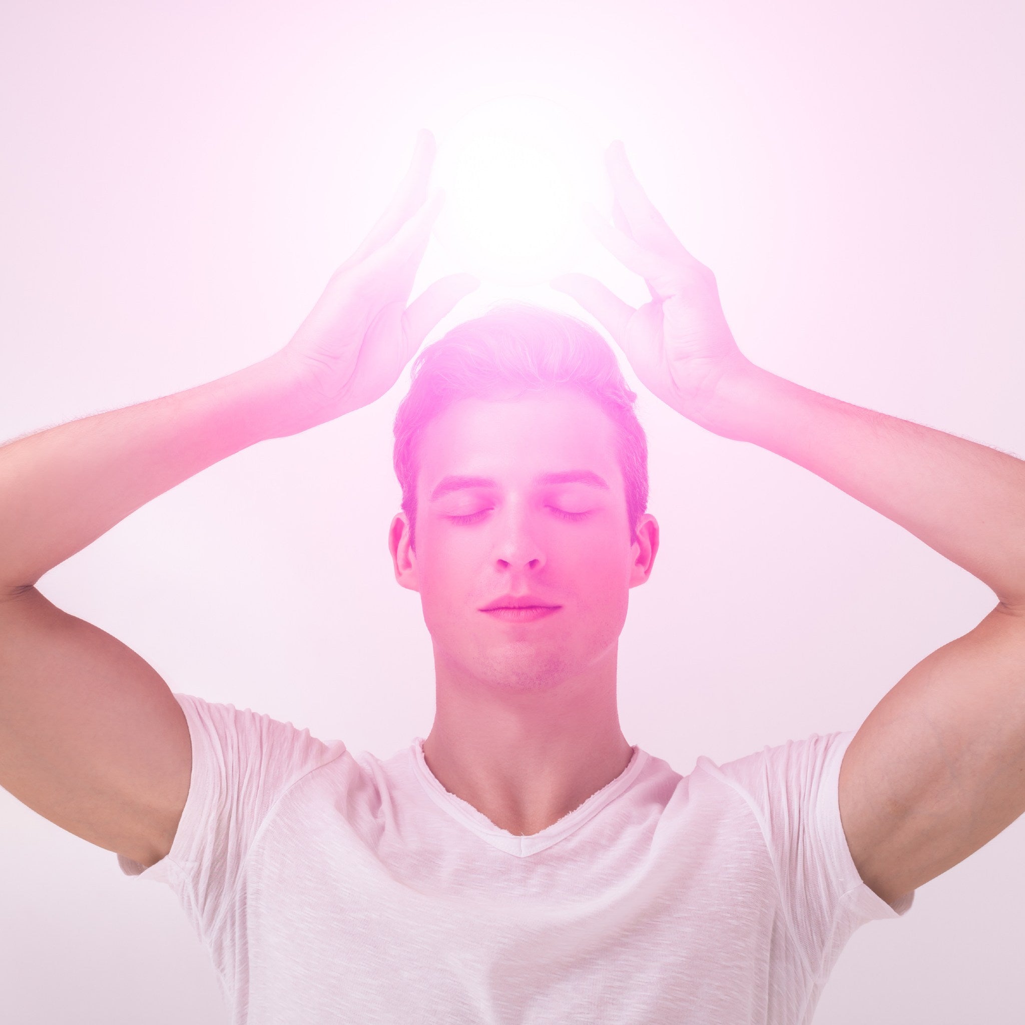 A young man with eyes closed and hands raised above his head, channeling an intense, bright white-pink light that glows above his crown, symbolising connection to higher consciousness.