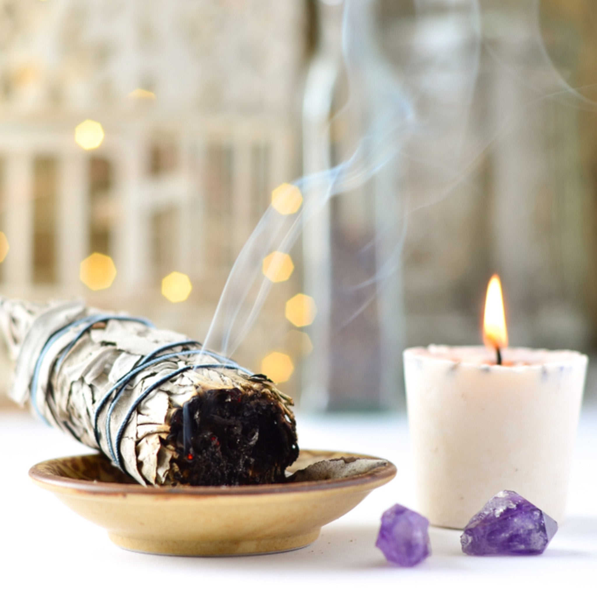Sage bundle on a dish with smoke, a lit candle, and amethyst crystals on a blurred background symbolising removing negative energy and space clearing.