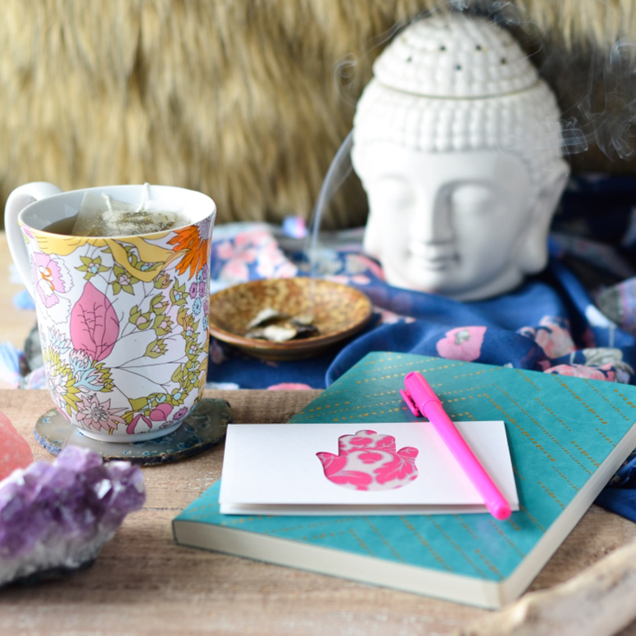 A cozy arrangement of a patterned mug with tea, a notebook with a pink pen, an amethyst crystal, and a white Buddha head incense burner emitting smoke.
