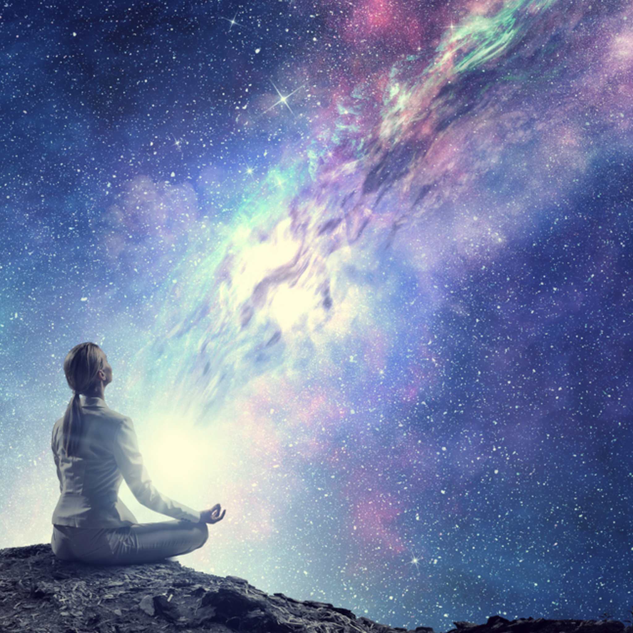 Silhouette of a figure sitting in a yoga or meditation pose on a rocky outcrop, looking up at a vast, bright, purple and blue cosmic galaxy.