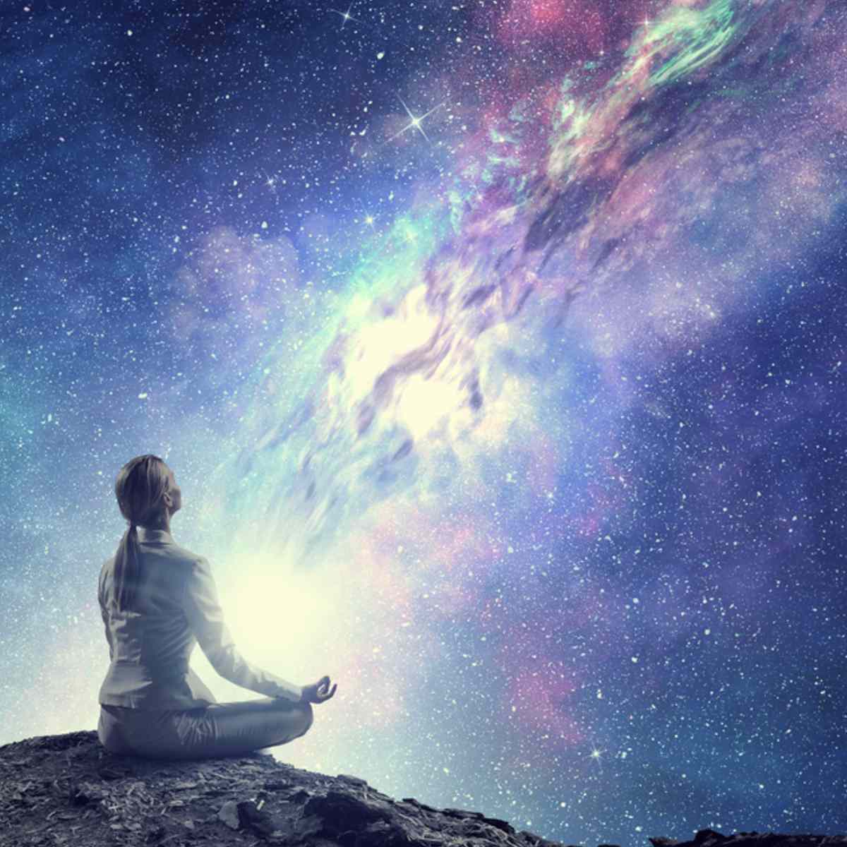 Silhouette of a figure sitting in meditation on a rocky outcrop, looking up at a vast, bright, purple and blue cosmic galaxy.
