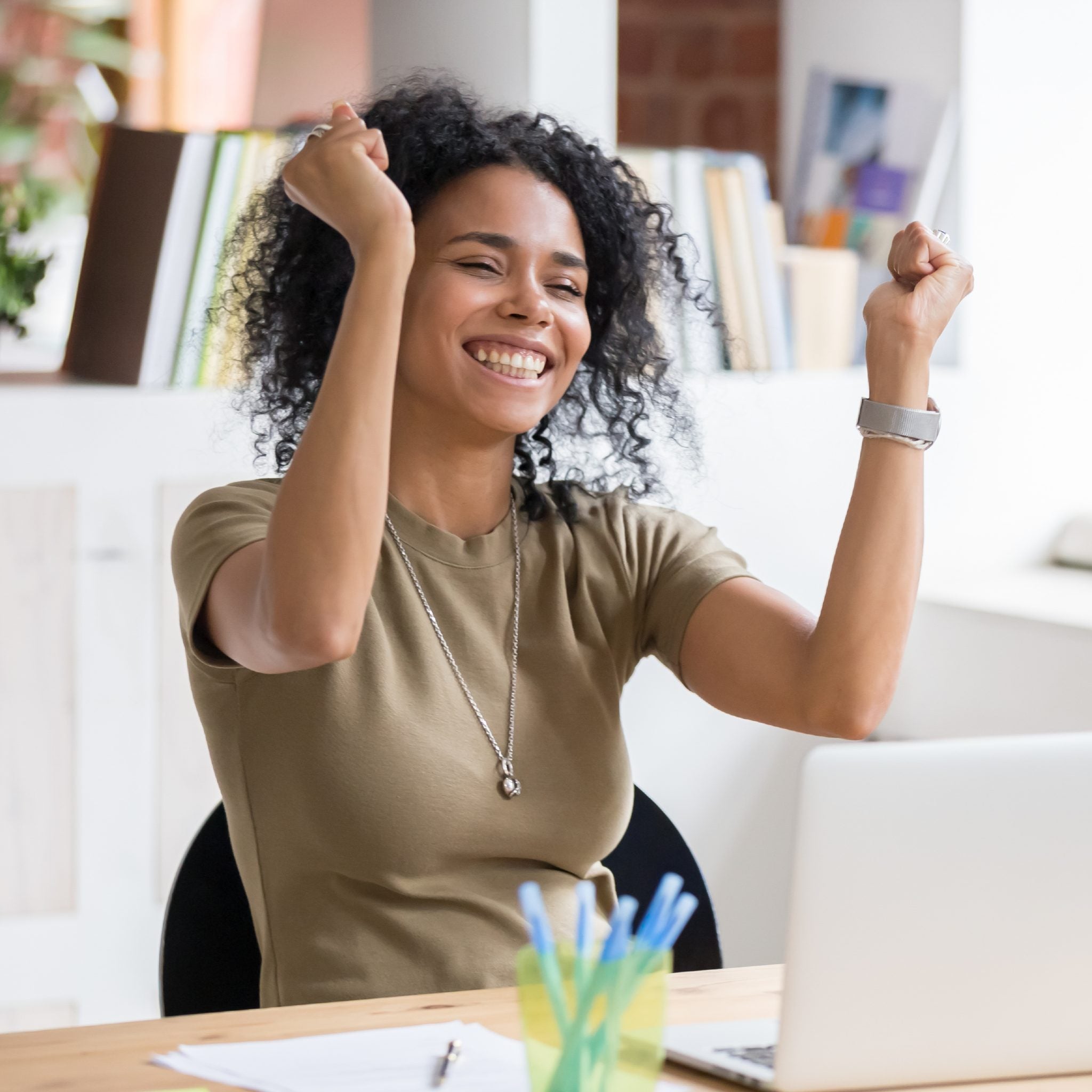 A photograph of a young woman with curly dark hair sitting at a desk in a bright office, smiling broadly at the camera with her hands raised in fists of triumph. She is celebrating a victory at her laptop, symbolising successful business.