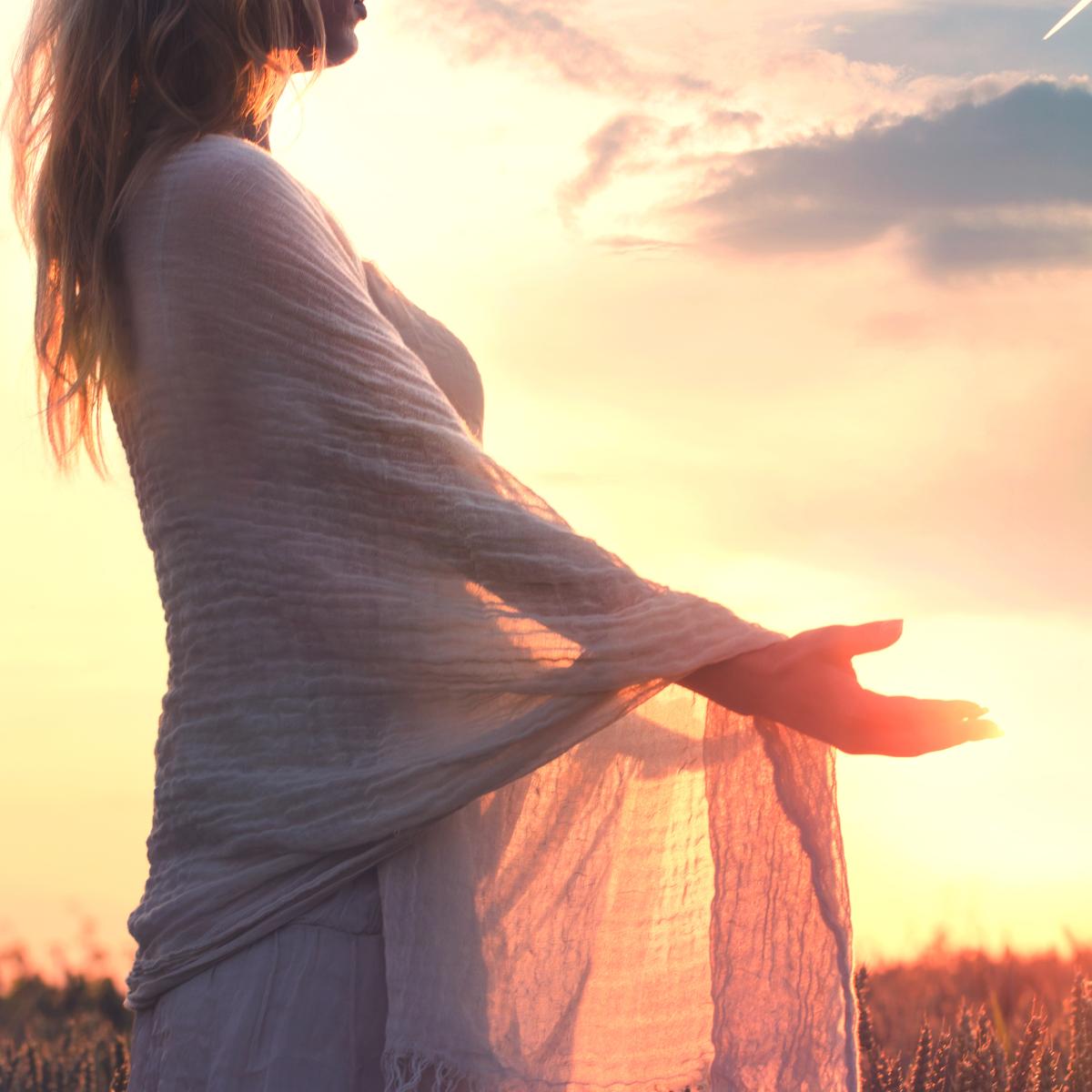 A woman in a white, flowing scarf stands in a field with her arms open at sunset, representing the peaceful surrender rite.