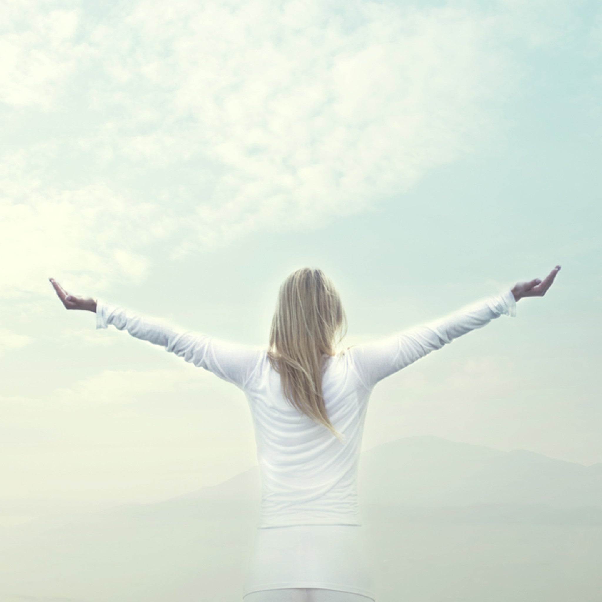 Synchronicity Rite: a person in white clothing standing outdoors with arms wide open, facing a vast, misty landscape under a soft, luminous sky.