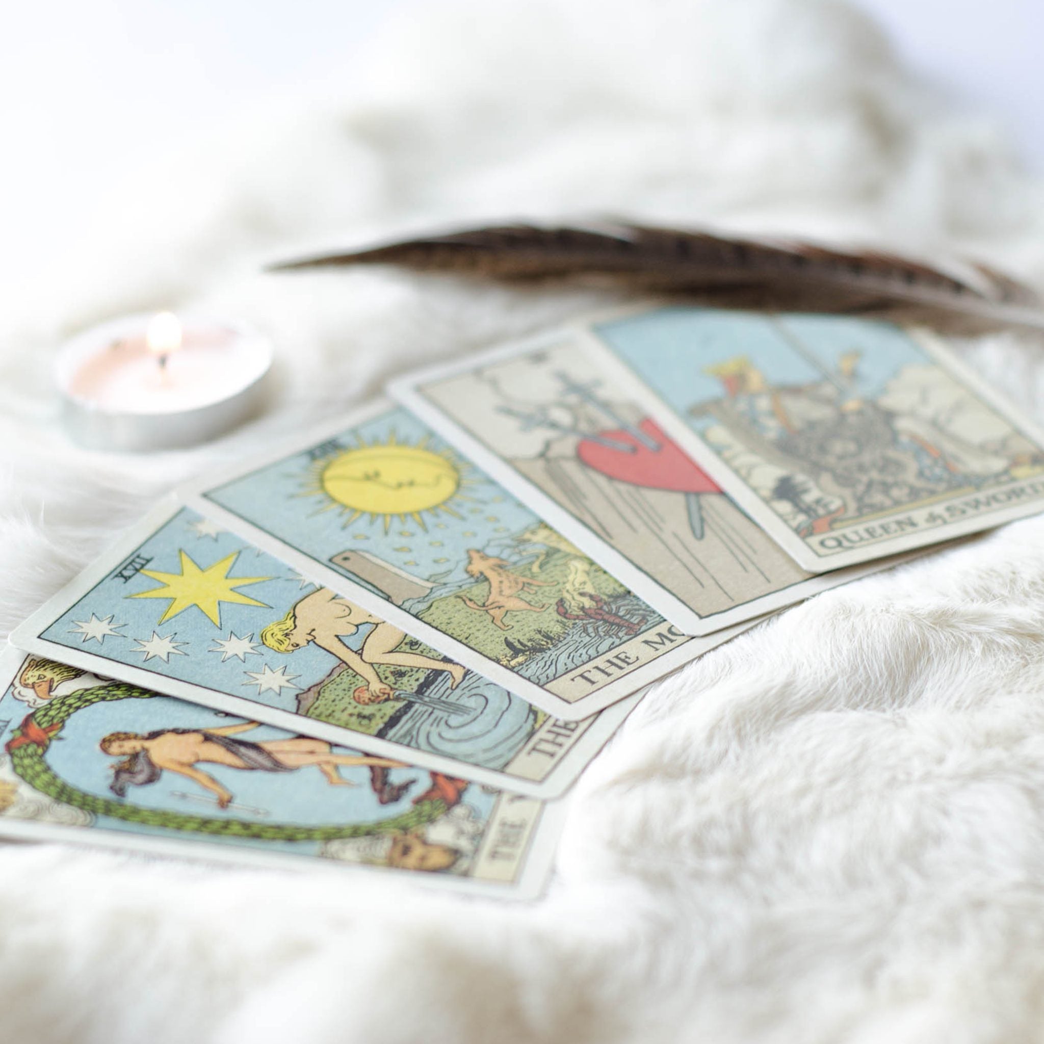 A close-up, soft-focus photograph of four classic Tarot cards laid out on a white, fluffy rug or throw, alongside a flickering tea light and a feather. The visible cards include The Star and The World, representing the focus on intuition and clarity in the Tarot reader path alignment dedication.