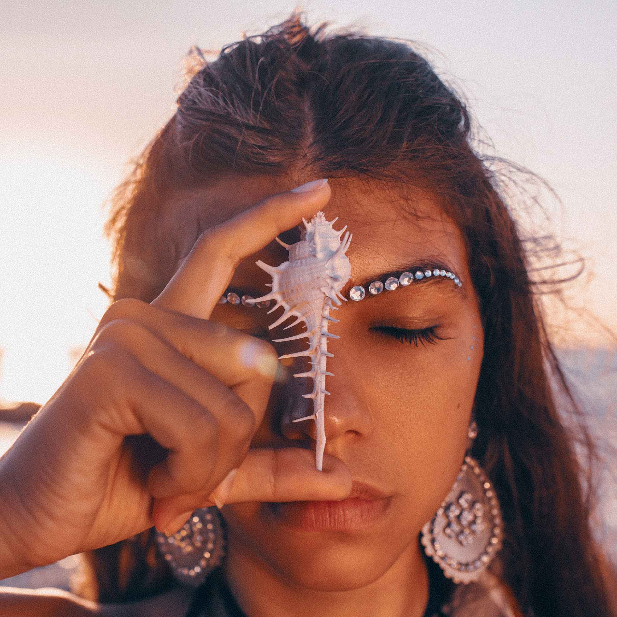 A close-up portrait of a woman with her eyes closed and a strip of glittering Rhinestones or jewels across her forehead. She is holding a long, spiky white conch shell vertically over her face and third eye, with the tip resting on her nose, against a bright, hazy, sunset background.