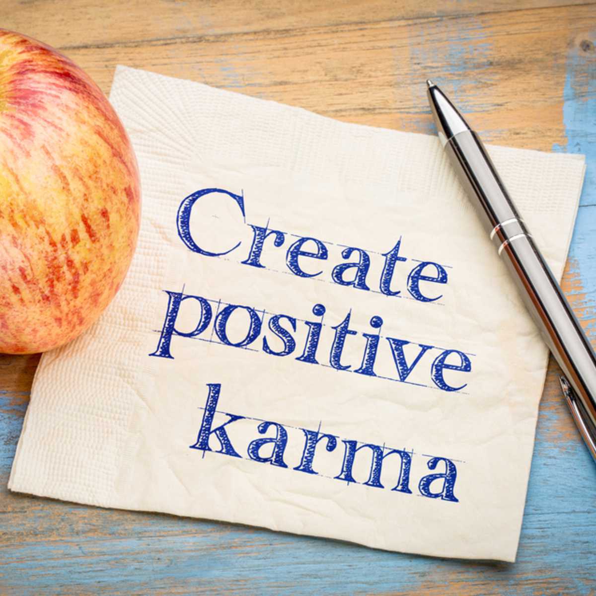 Napkin with 'Create positive karma' text, apple, and pen on a wooden surface.