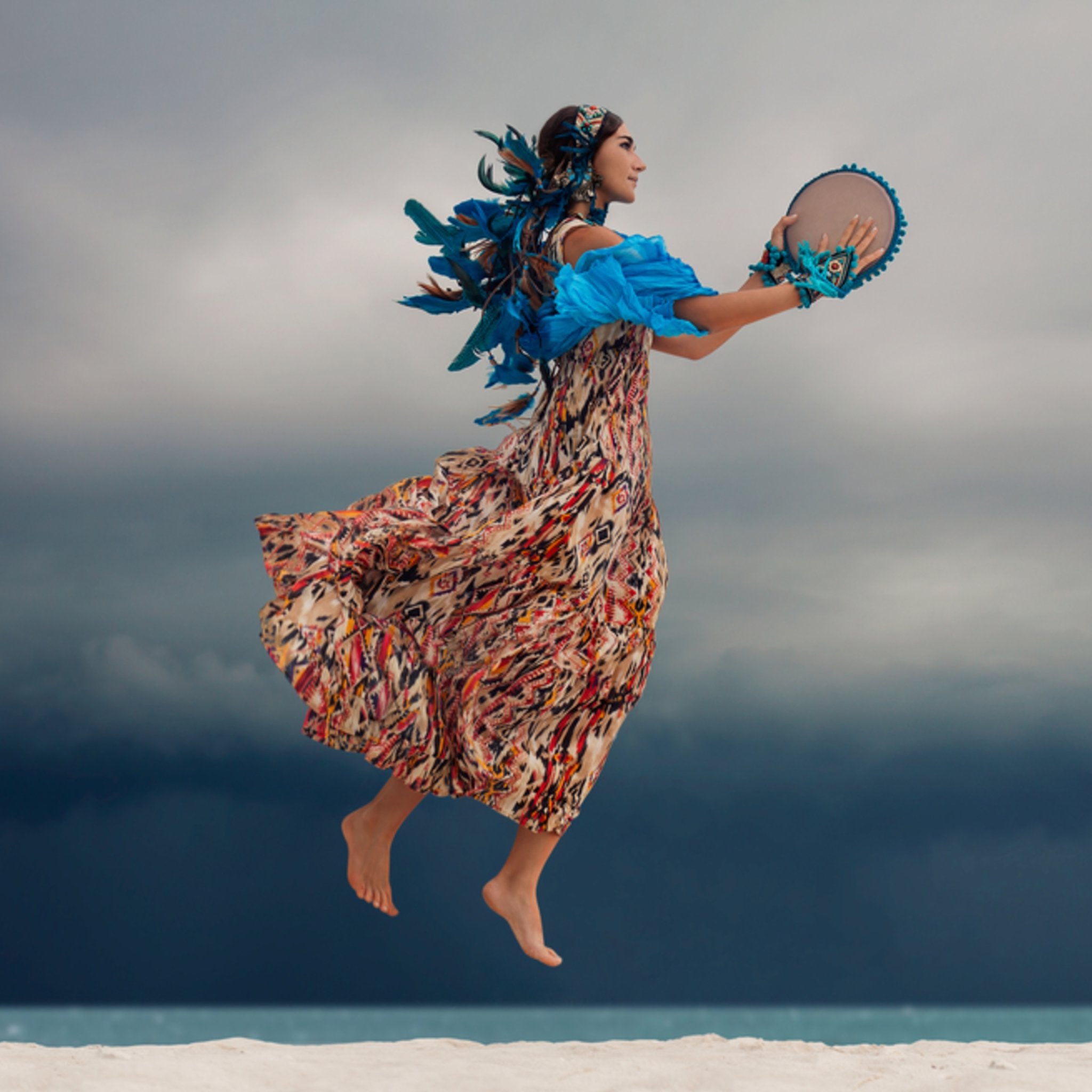 A barefoot woman in a colourful, patterned dress and elaborate blue feather and beaded headdress jumps high above white sand, holding a small round drum. Her flowing garments and feathers are accentuated by the mid-air pose. The background is a dramatic dark grey and stormy blue sky over a distant body of water, illustrating a shamanic soul retrieval concept.