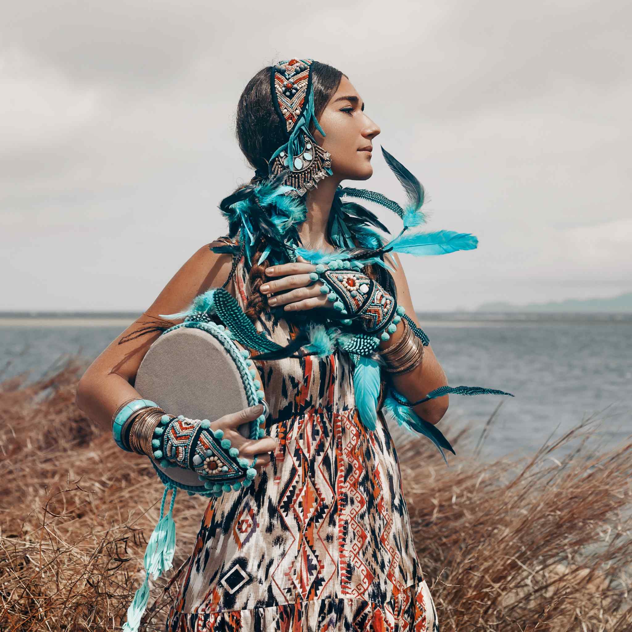 A woman outdoors, wearing a long patterned dress and heavy, turquoise feather and beaded jewellery, including a headband and cuffs. She holds a small, round hand drum to her chest and looks off to the right. This image represents a shamanic warrior concept. She stands in dry grass with a body of water and distant land in the background.
