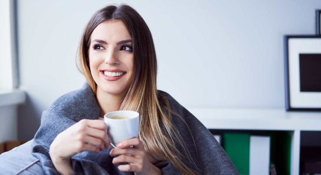 Woman holding a mug with a cozy blanket draped over her shoulders in a room.
