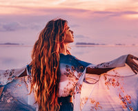 Woman with long hair standing by a body of water at sunset, wearing a floral dress.