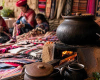 A clay pot cooking over a stove hearth, with Andean women in traditional red hats weaving vibrant textiles behind.