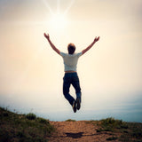 A photograph of a person in a white t-shirt and dark jeans jumping high in the air with their arms outstretched, standing on a dirt path atop a hill. The background is a bright, overexposed sky with a radiating sunburst, symbolising the uplifting energy of the 5555 angel number.
