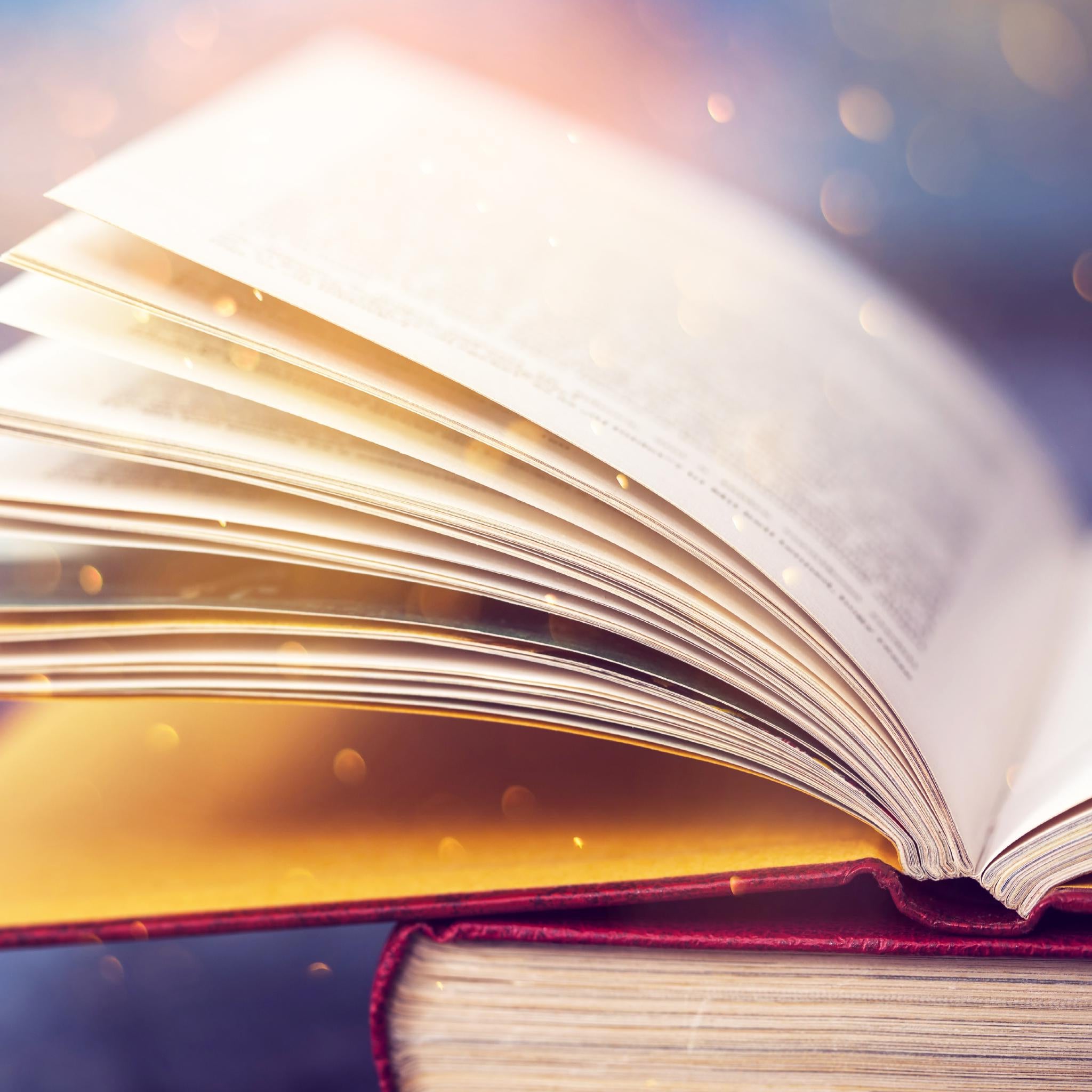 Close-up of a large, opened book with pages gently fanned open, illuminated by a warm, ethereal light and golden bokeh sparkles.
