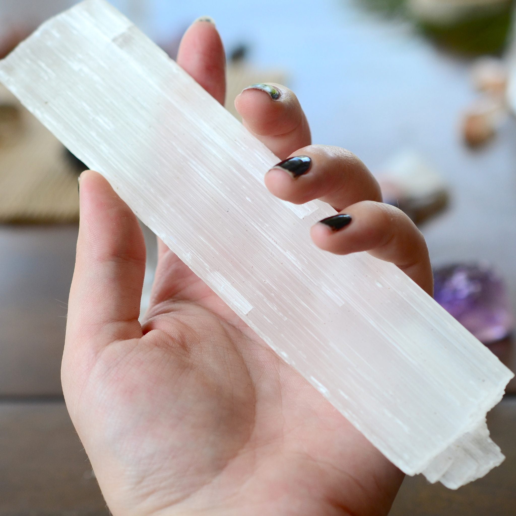 A close-up photograph of a person's hand gently holding a long, rectangular piece of white selenite crystal, with black painted fingernails. The image focuses on the clear crystalline structure against a soft, blurred background of natural light and subtle colours, representing angelic selenite protection.