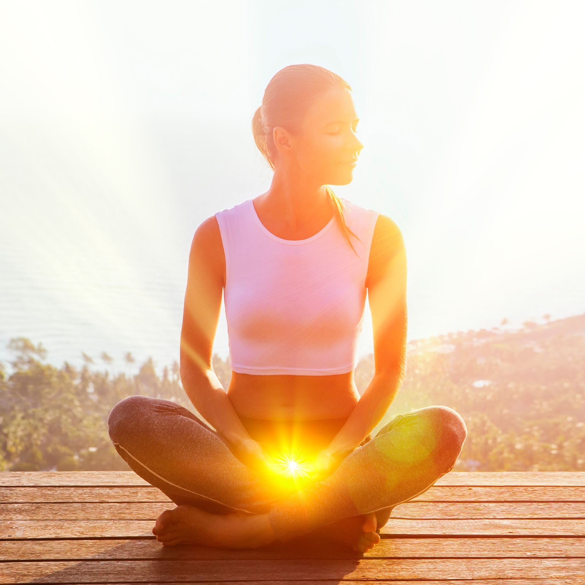 A woman sits in a cross-legged yoga pose on a wooden deck overlooking a sunny, tropical landscape, with an intense glowing orange-yellow light emanating from her lap/root chakra area.