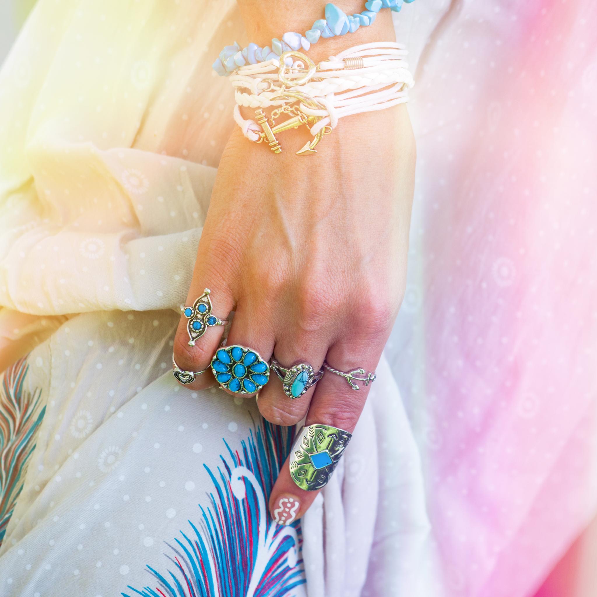 A close-up of a person's hand adorned with several silver rings featuring turquoise and blue stones, including a large flower ring. The arm is decorated with stacked bracelets, including white leather cords and blue beads, all set against a soft, sunlit background of flowing light-coloured fabric.