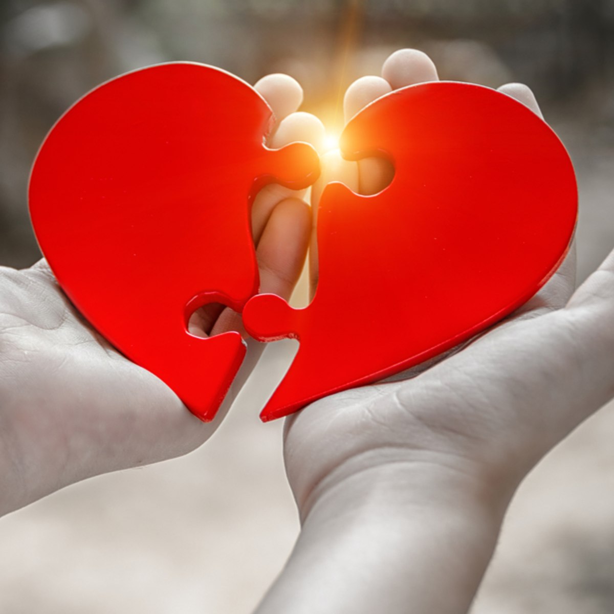 Two hands holding together two red pieces of a heart shape, with warm light shining between them.