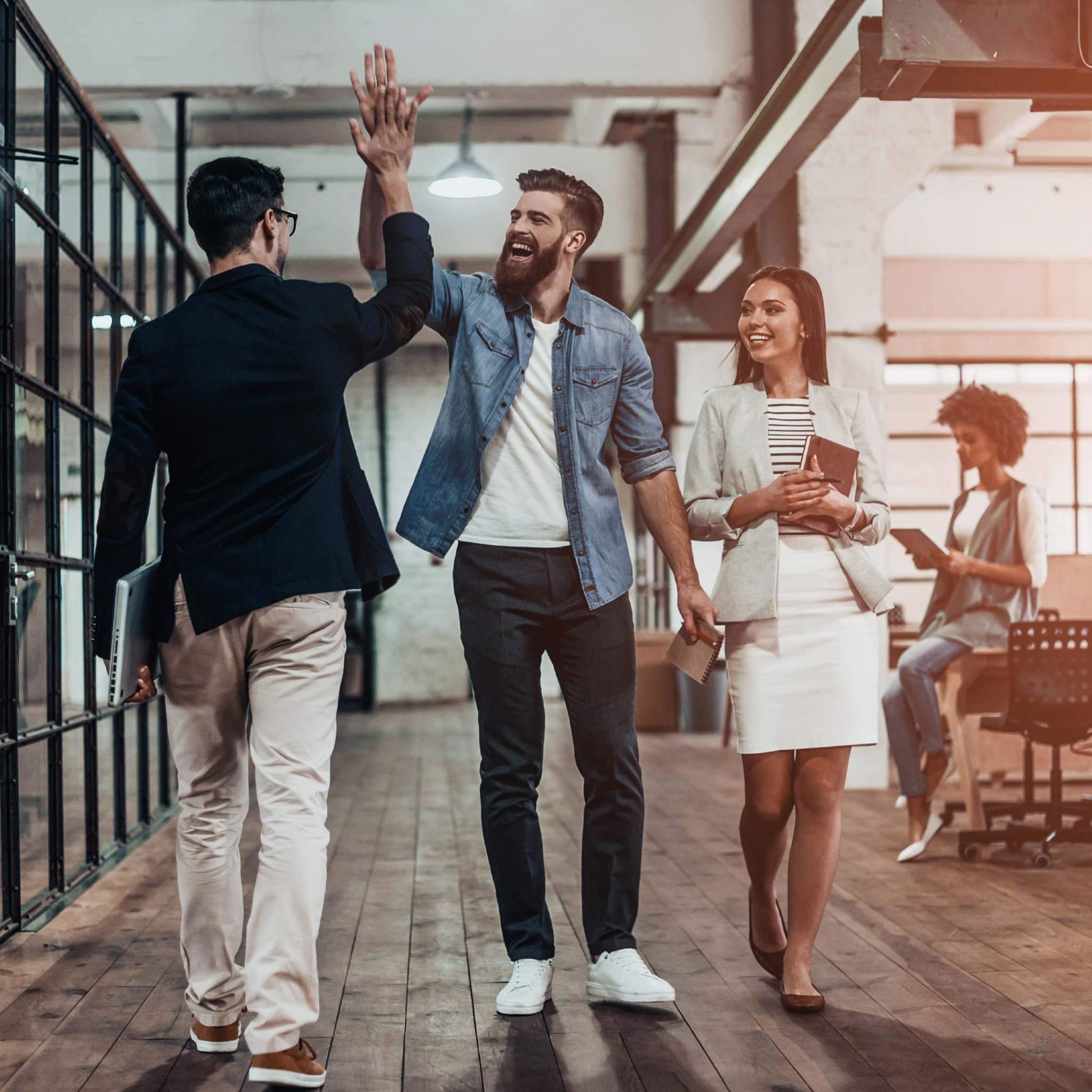 A photograph of two male colleagues walking down an open-plan office corridor with wooden floors, giving each other a high five and laughing. A female colleague smiles as she walks alongside them, while another colleague works in the background, symbolising a positive atmosphere following a business space clearing.