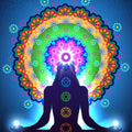 A silhouette of a meditating figure against a bright blue background, with the seven main chakras glowing and a large, vibrantly coloured mandala centred around the glowing Crown Chakra.