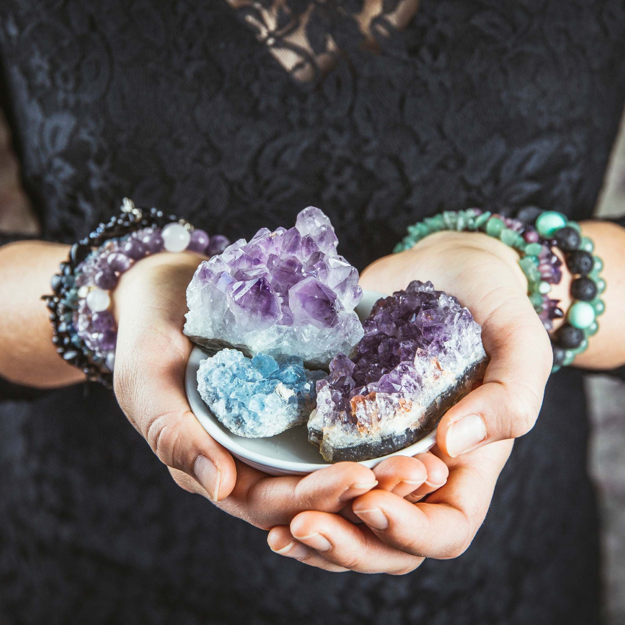 A close-up photograph of a person's hands, wearing several beaded bracelets, holding a small white dish filled with raw, purple amethyst and blue celestite crystals. The hands are gently cupped over the bowl against a dark background, symbolising the use of gemstones in the crystal healer path alignment dedication.