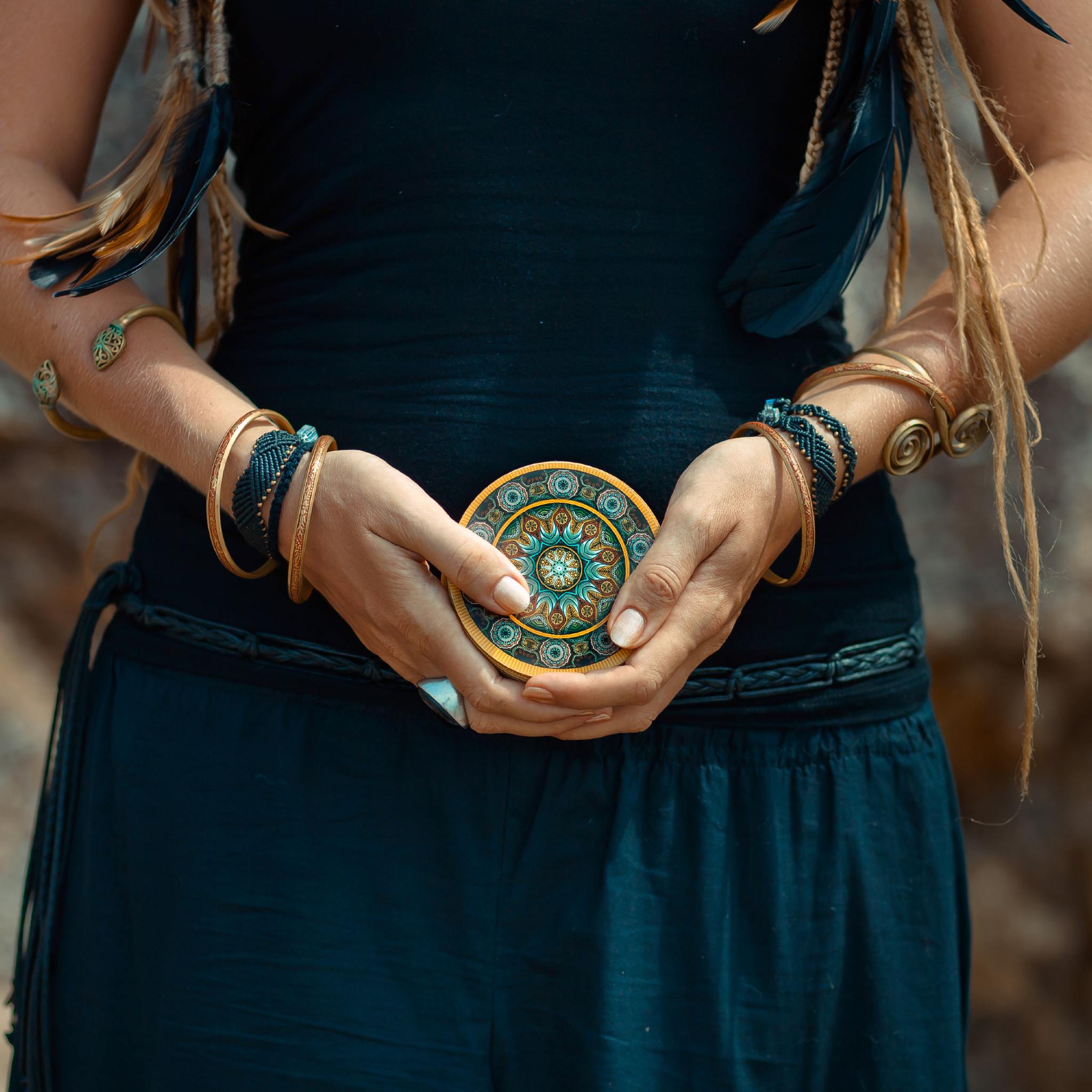 A shamanic woman with dreadlocks and tribal jewellery holding a round, detailed, colourful mandala symbol over her lower abdomen.