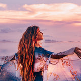 A woman with long, flowing hair and a blue patterned robe, arms outstretched toward a pink and purple sunset over calm water, representing dynamic Abundantia connections.