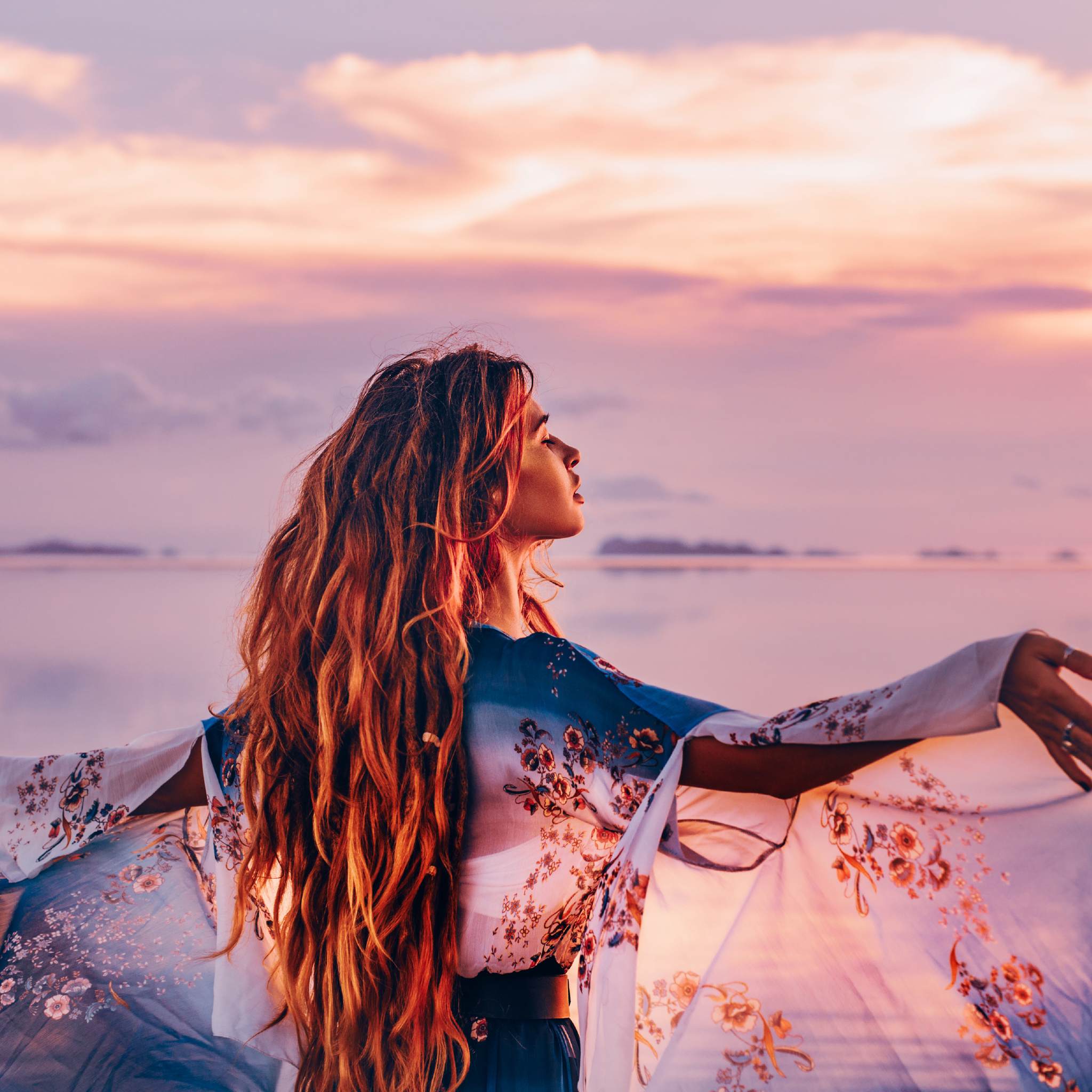 A woman with long, flowing hair and a blue patterned robe, arms outstretched toward a pink and purple sunset over calm water, representing dynamic Abundantia connections.