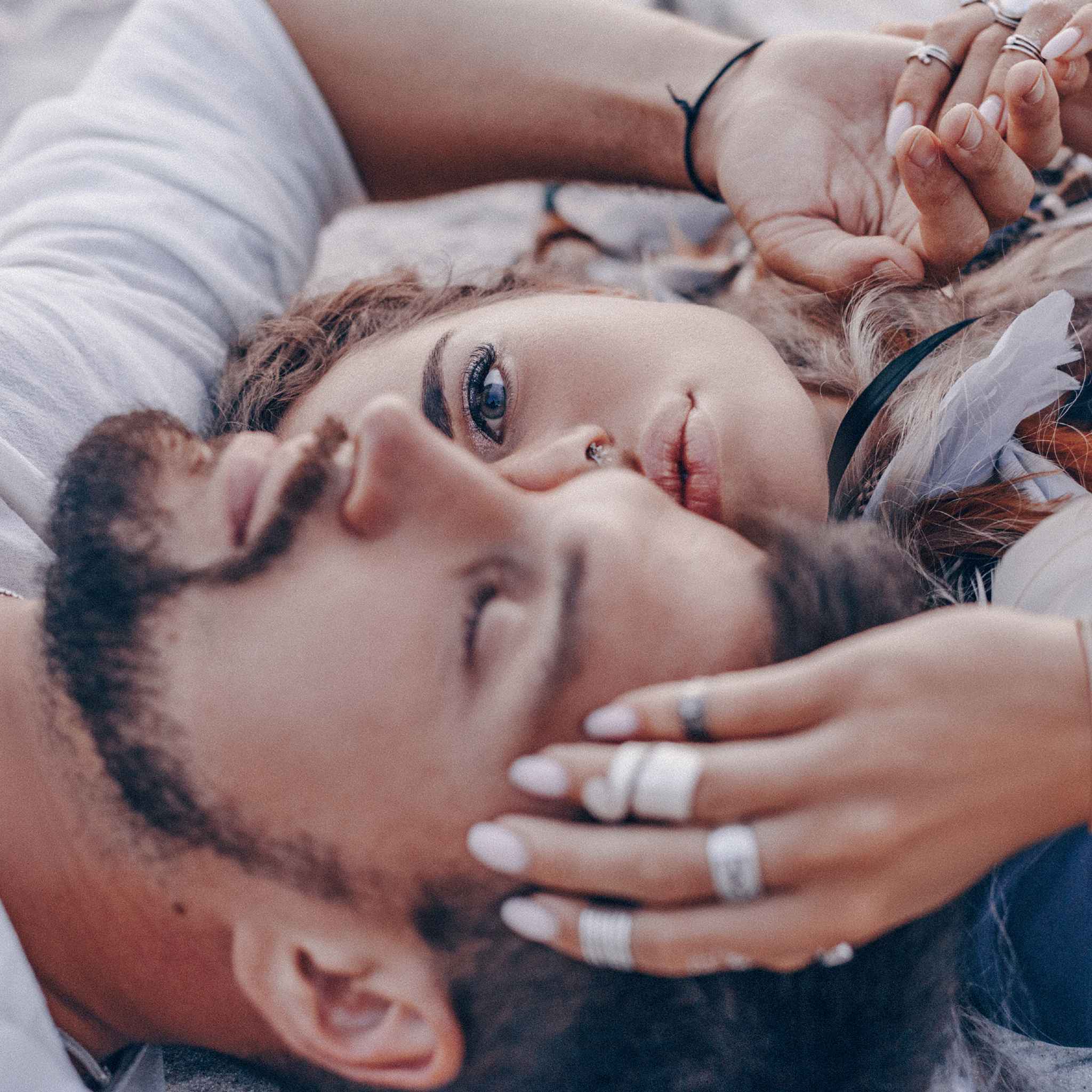 A close-up, romantic photo of a couple lying down, where the woman is looking directly at the camera with intense blue eyes and one hand resting gently on the man's head, both adorned with rings and soft-toned accessories.