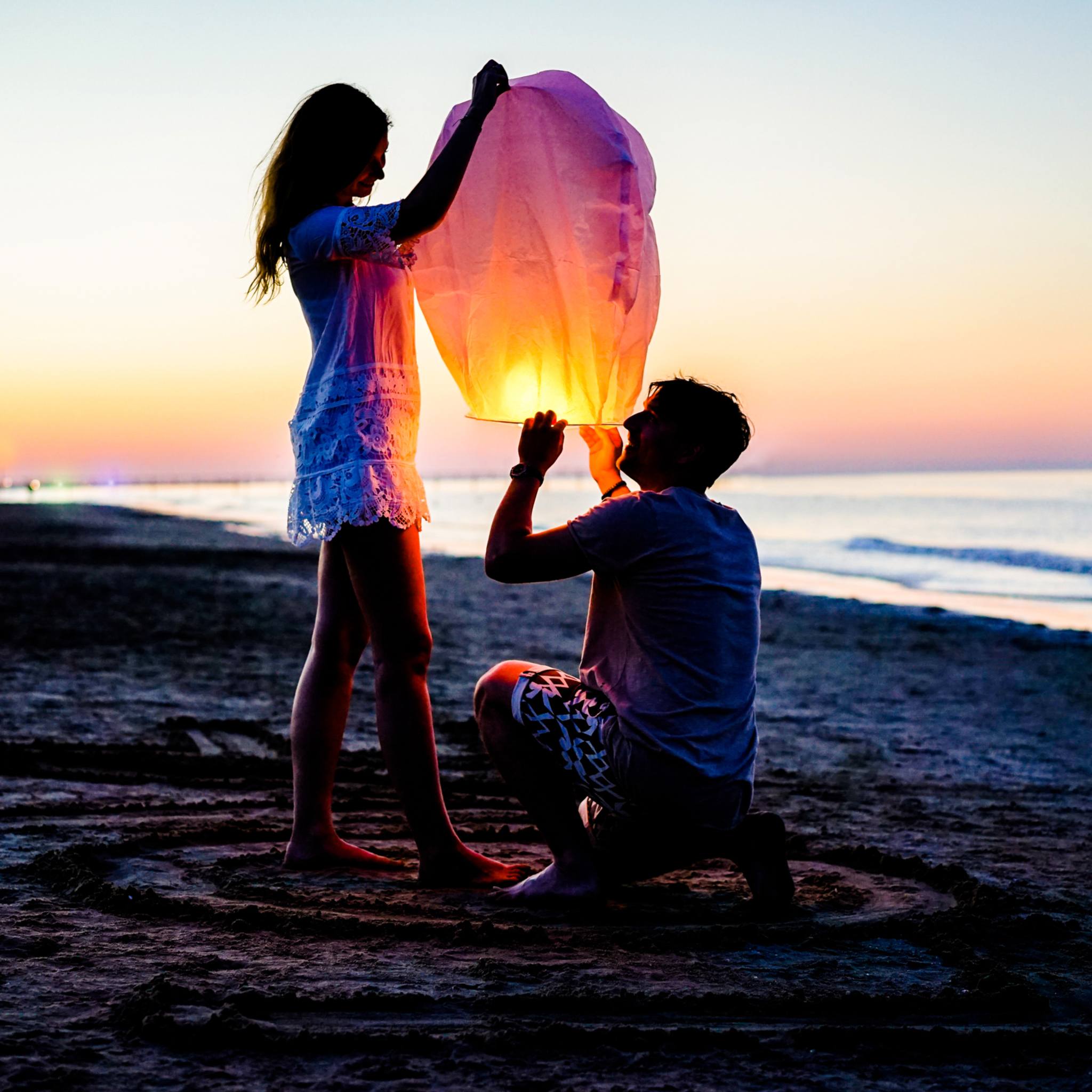A couple on a beach at sunset, lighting a glowing paper lantern together, representing eros connections.