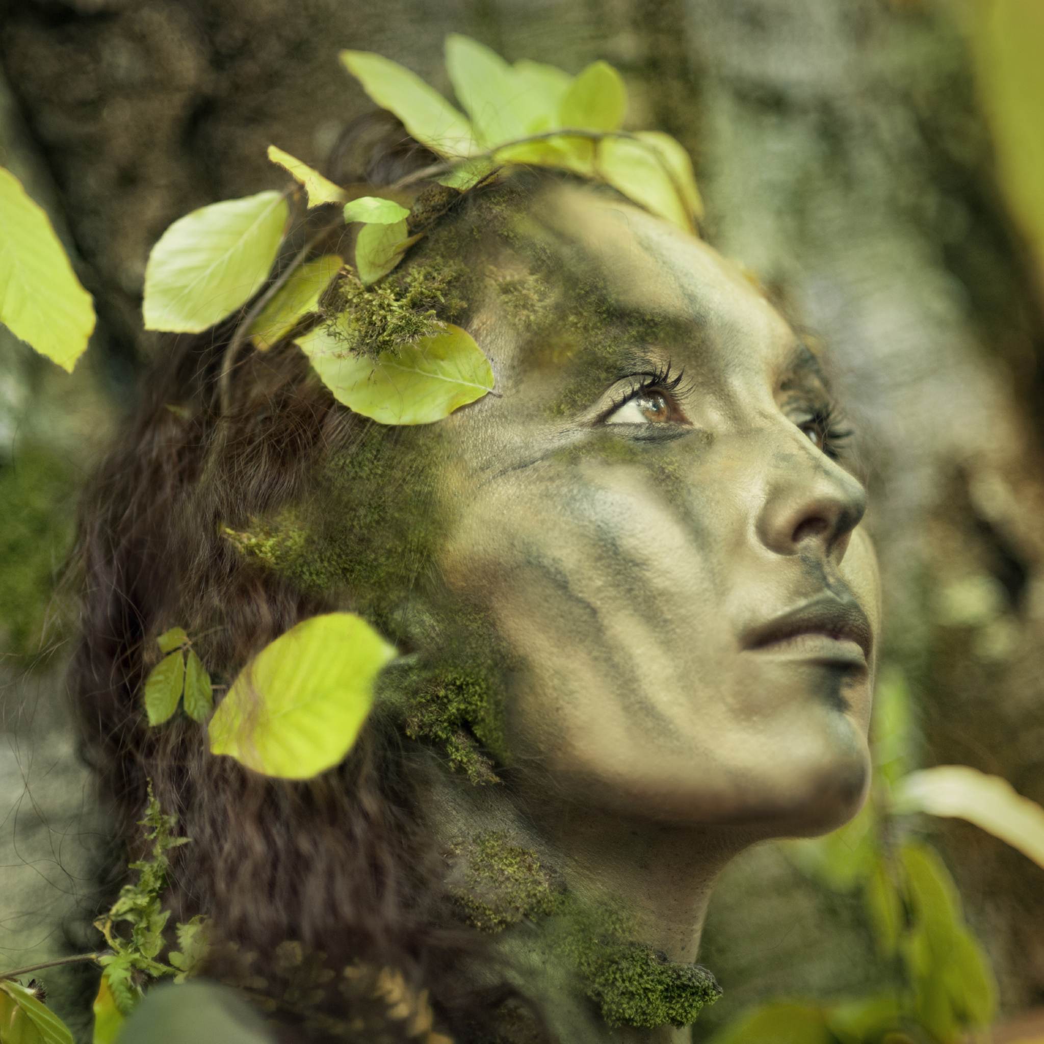 A beautiful woman with her face painted in camouflage green and moss, wearing leaves in her hair, looking upwards, representing Green Woman connections.