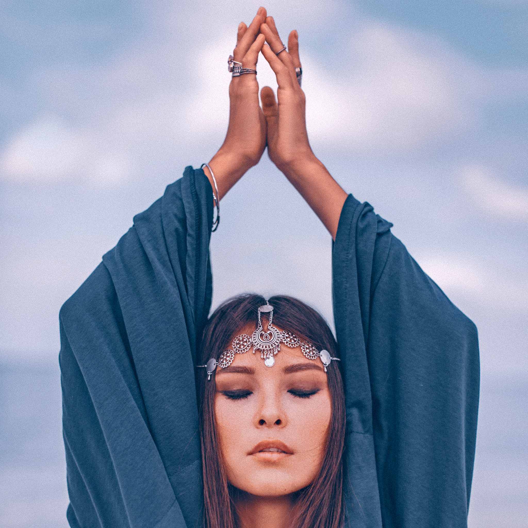 A serene woman with her eyes closed, wearing a silver forehead jewellery piece, raises her hands in prayer or offering above her head, draped in a deep blue-grey shawl or robe against a pale, cloudy sky.