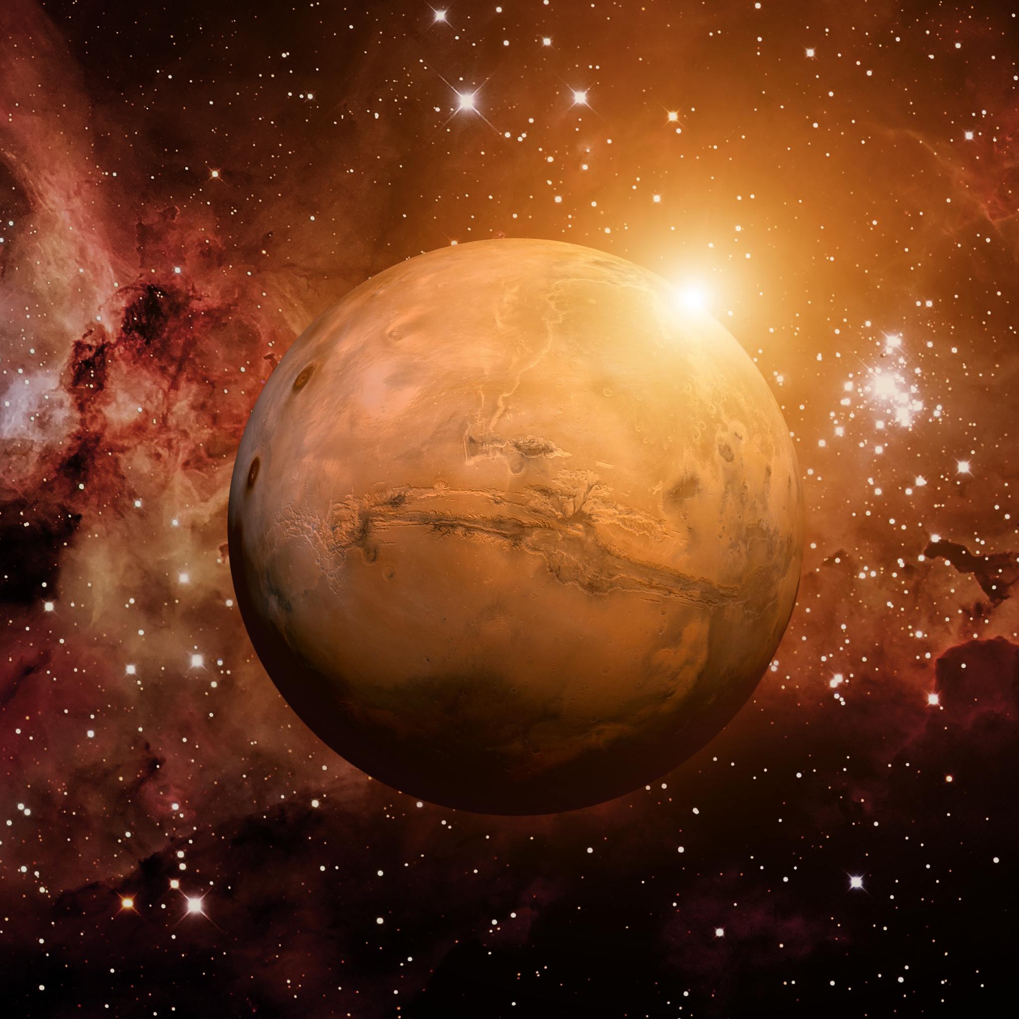 A detailed, high-resolution image of the planet Mars, showing its characteristic reddish-orange surface, illuminated by a bright sunflare near the top right. The planet is set against a dark space background filled with distant stars and a swirling red and orange nebula, symbolising the intense Mars energy connections.