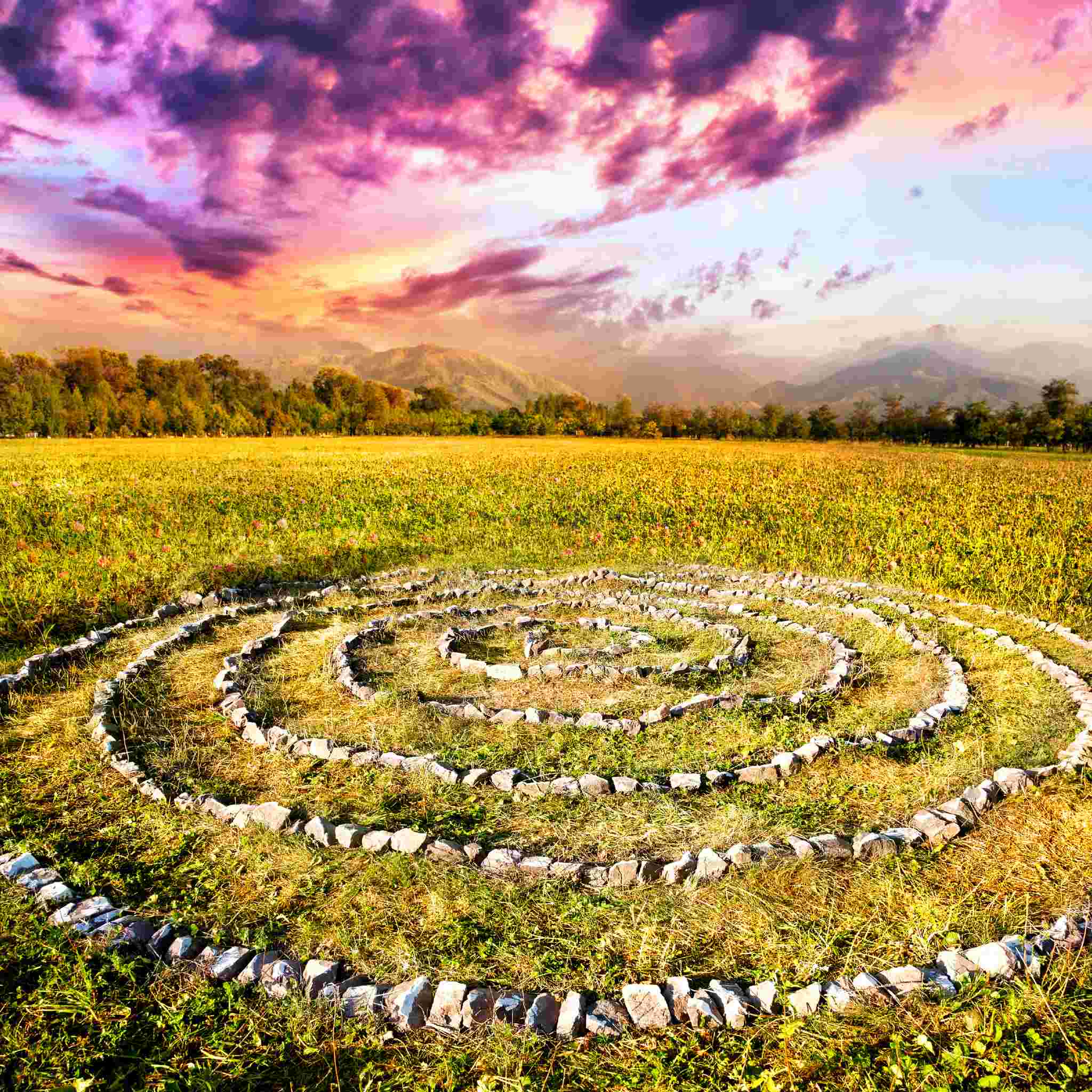 An outdoor photograph of a spiralling arrangement of light-coloured stones laid out on a field of green and yellow grass, forming a medicine wheel or labyrinth. The background features a vibrant sunset with pink and purple clouds over distant hills and forestt.