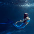 A woman with long red hair and a shimmery blue mermaid tail swimming gracefully underwater.
