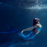 A woman with long red hair and a shimmery blue mermaid tail swimming gracefully underwater.