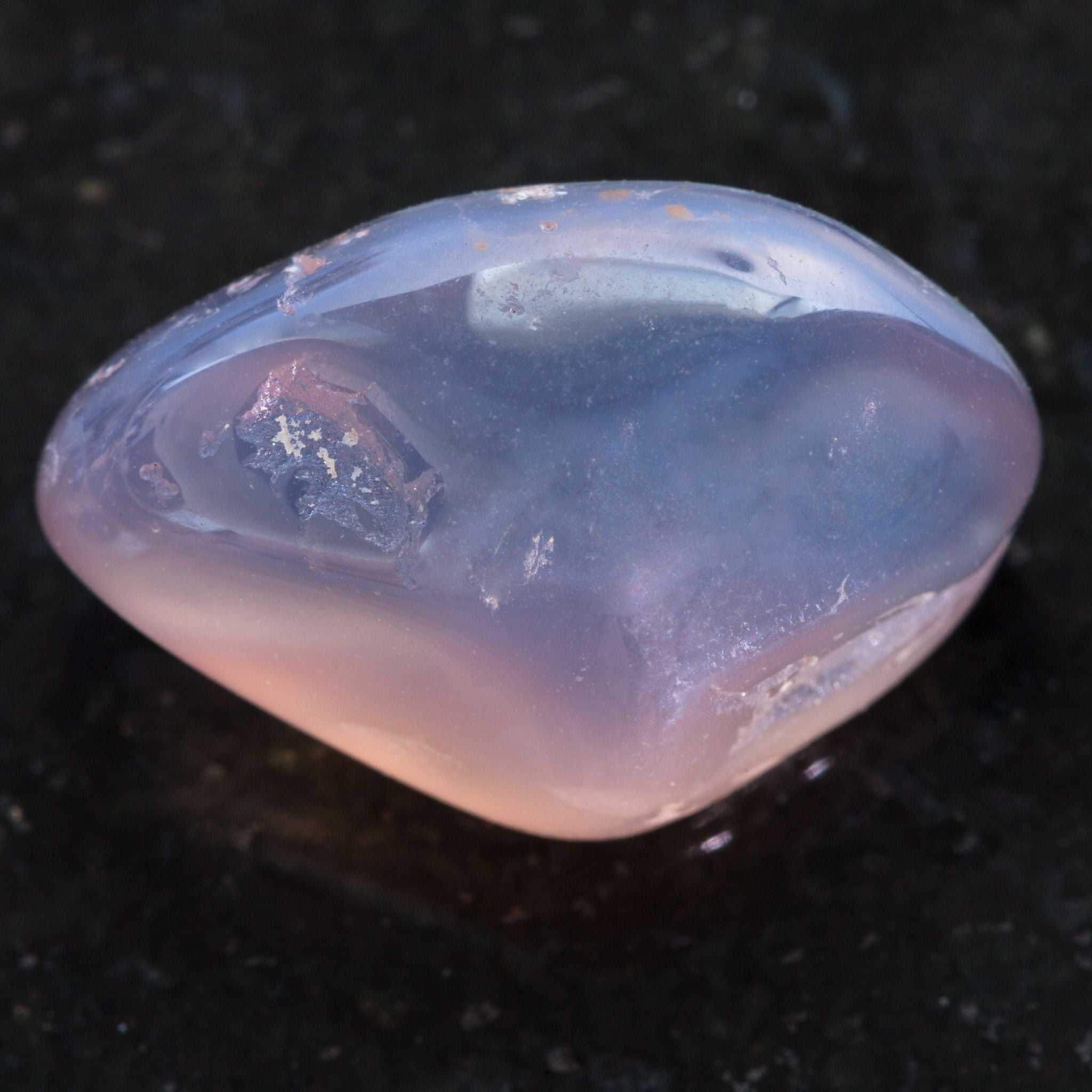 A macro close-up of a smooth, tumbled Moonstone crystal, showing its characteristic translucent, milky-white to pale-blue sheen and slight pink iridescence, set against a dark, speckled rock surface.