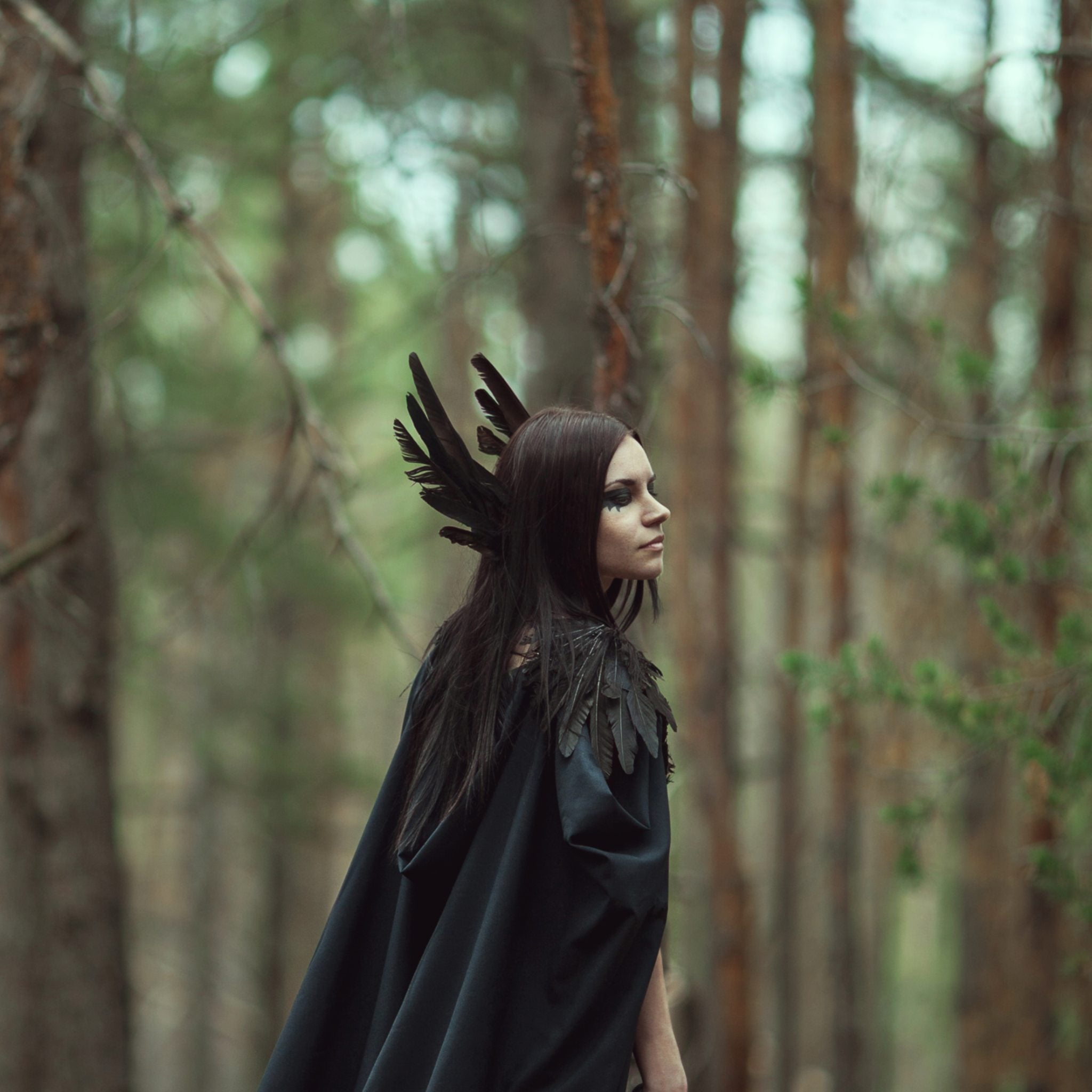 A woman with long black hair, wearing a dark cape, standing in a pine forest, representing Morrigan connections.