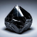 A close-up studio shot of a raw, faceted piece of black Obsidian, displaying a glassy texture and white fracture lines, brilliantly reflecting a studio light against a soft grey background.