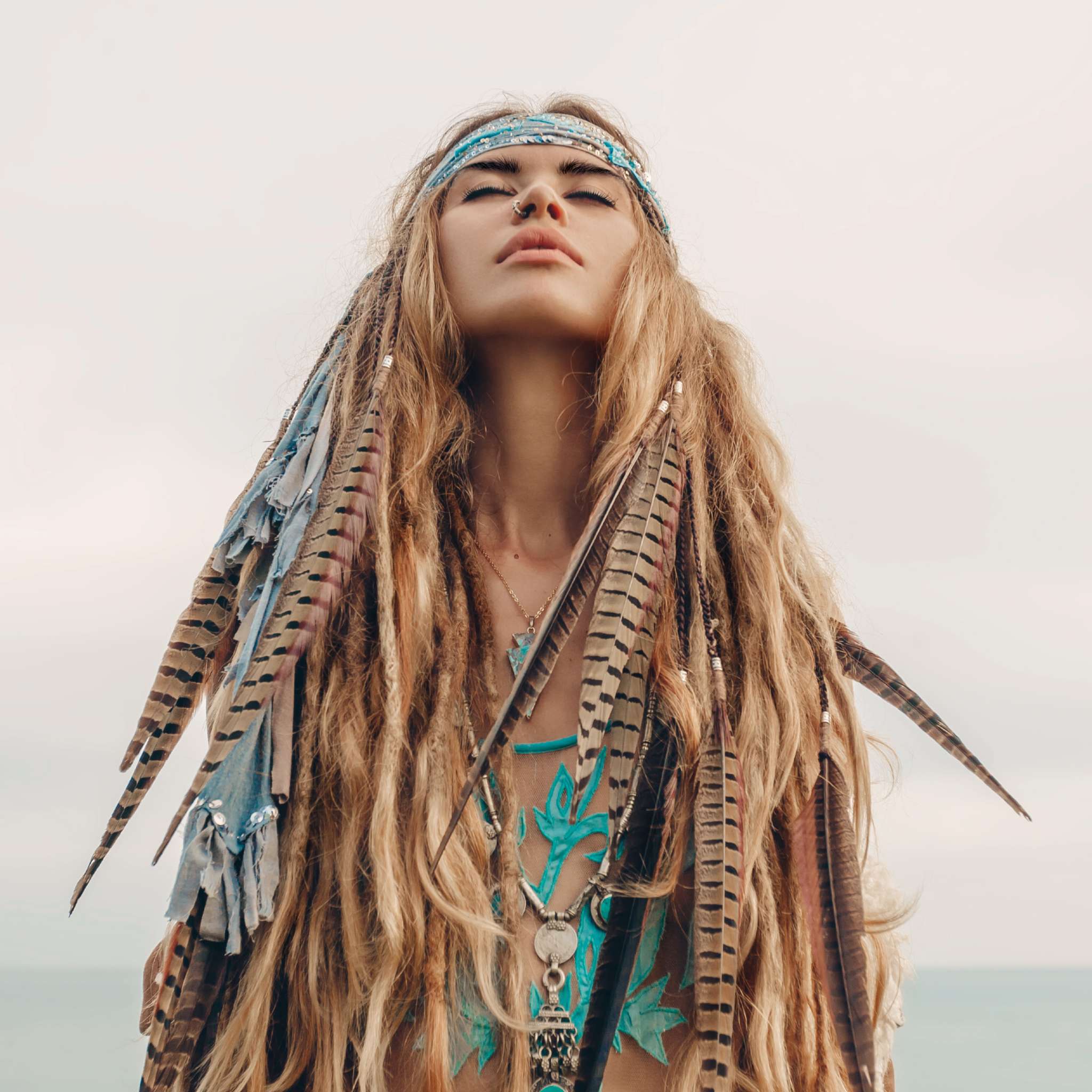 A close-up portrait of a woman with long, flowing blonde hair adorned with large feathers and blue cloth braids, wearing a decorative blue headband and a light nose ring. Her eyes are closed, and her face is lifted upwards against a soft, pale sky and sea background, signifying shamanic connections.