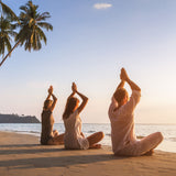 Three people practising yoga on a beach facing the sun at sunset with palm trees in the background.